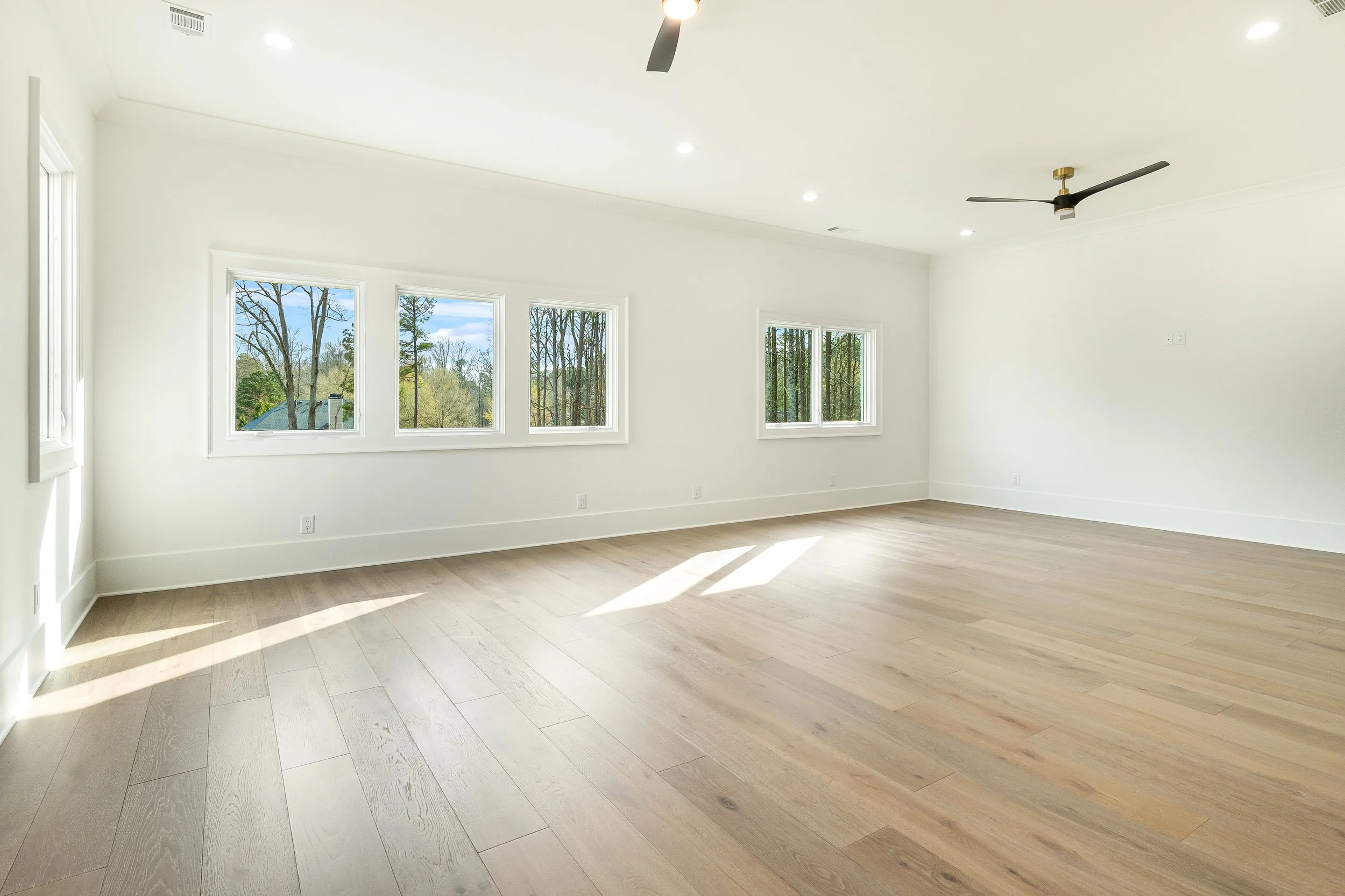 Empty white room with large windows, wooden flooring, ceiling fan, and recessed lighting.