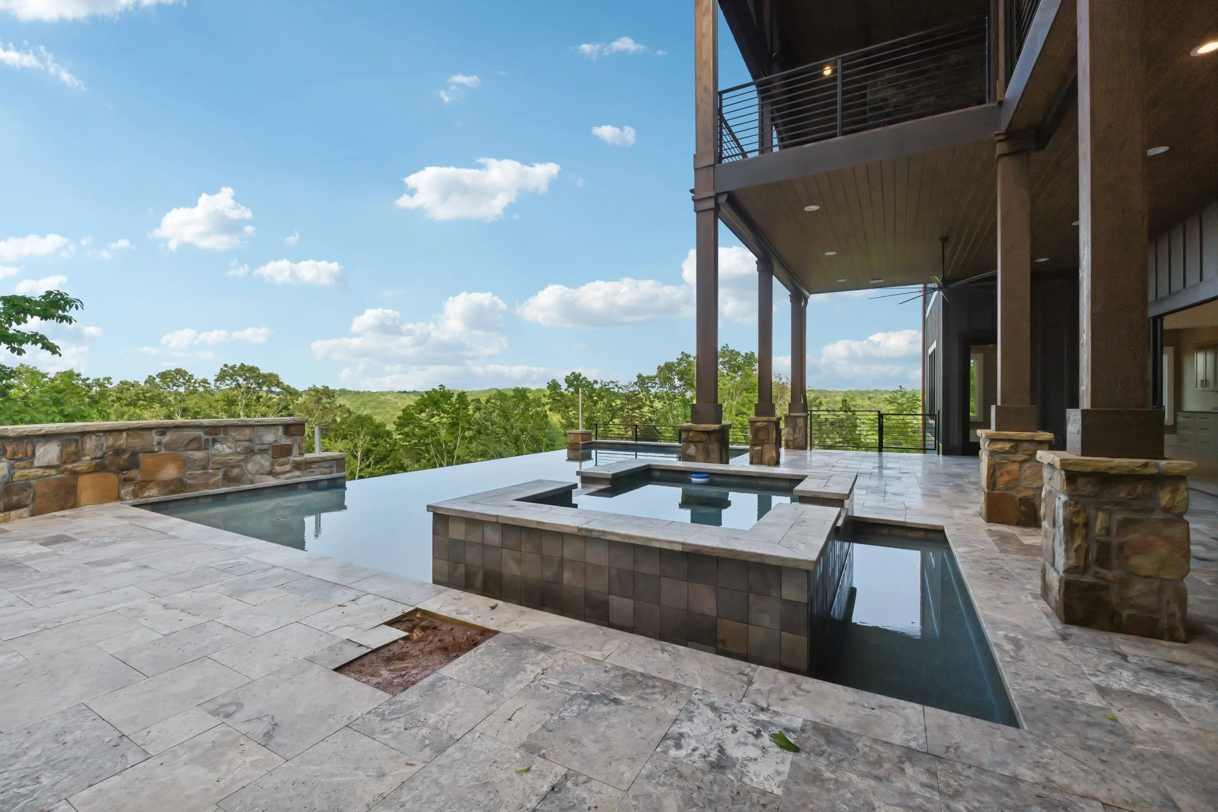 Modern house outdoor patio featuring a pool with a stone border, stone wall, wooden balcony, lush green trees, and a bright blue sky with white clouds.