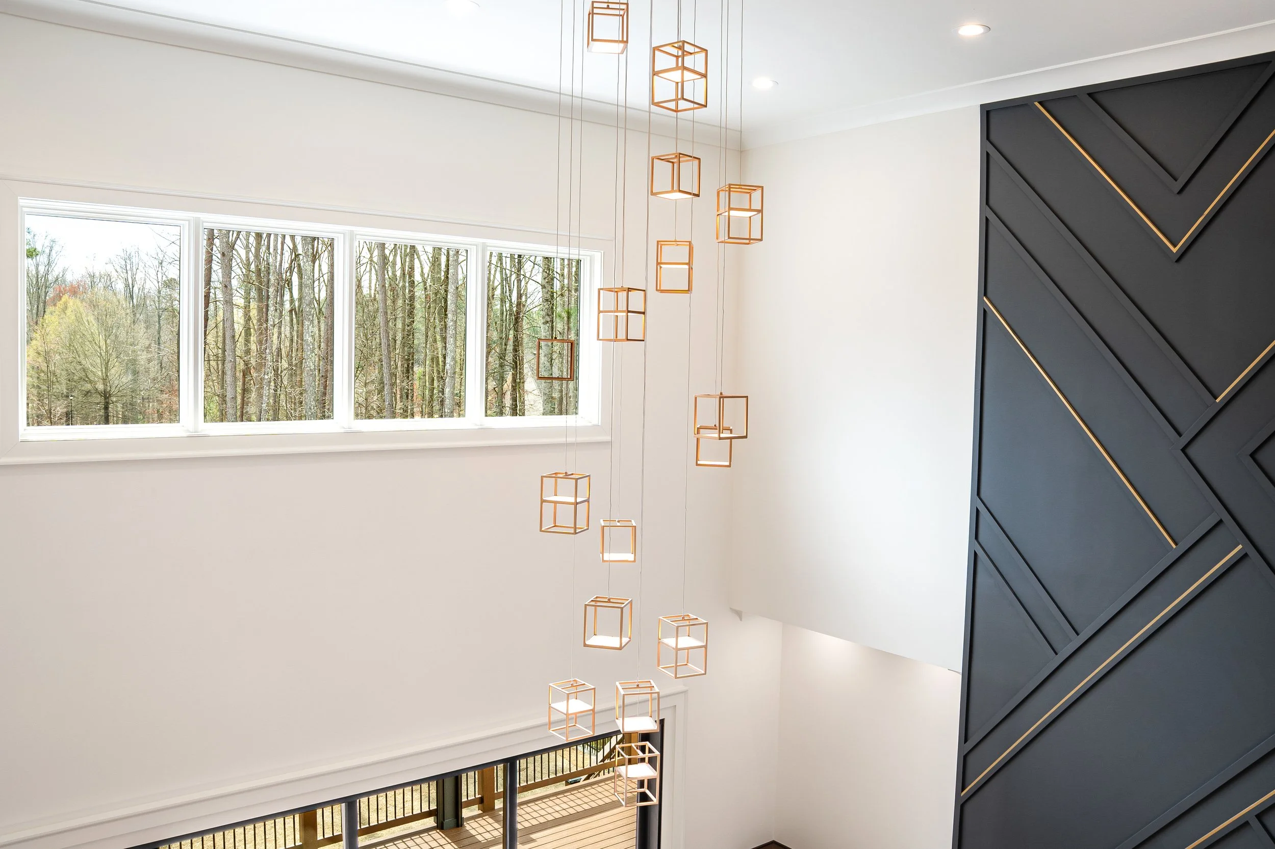 Interior view of a modern home with large windows, a black wall with geometric gold accents, and a hanging sculpture with multiple wooden cube frames.
