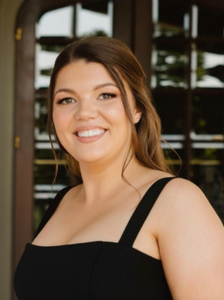 A young woman with long brown hair smiling, wearing a black sleeveless top, standing outdoors with a building and green trees in the background.