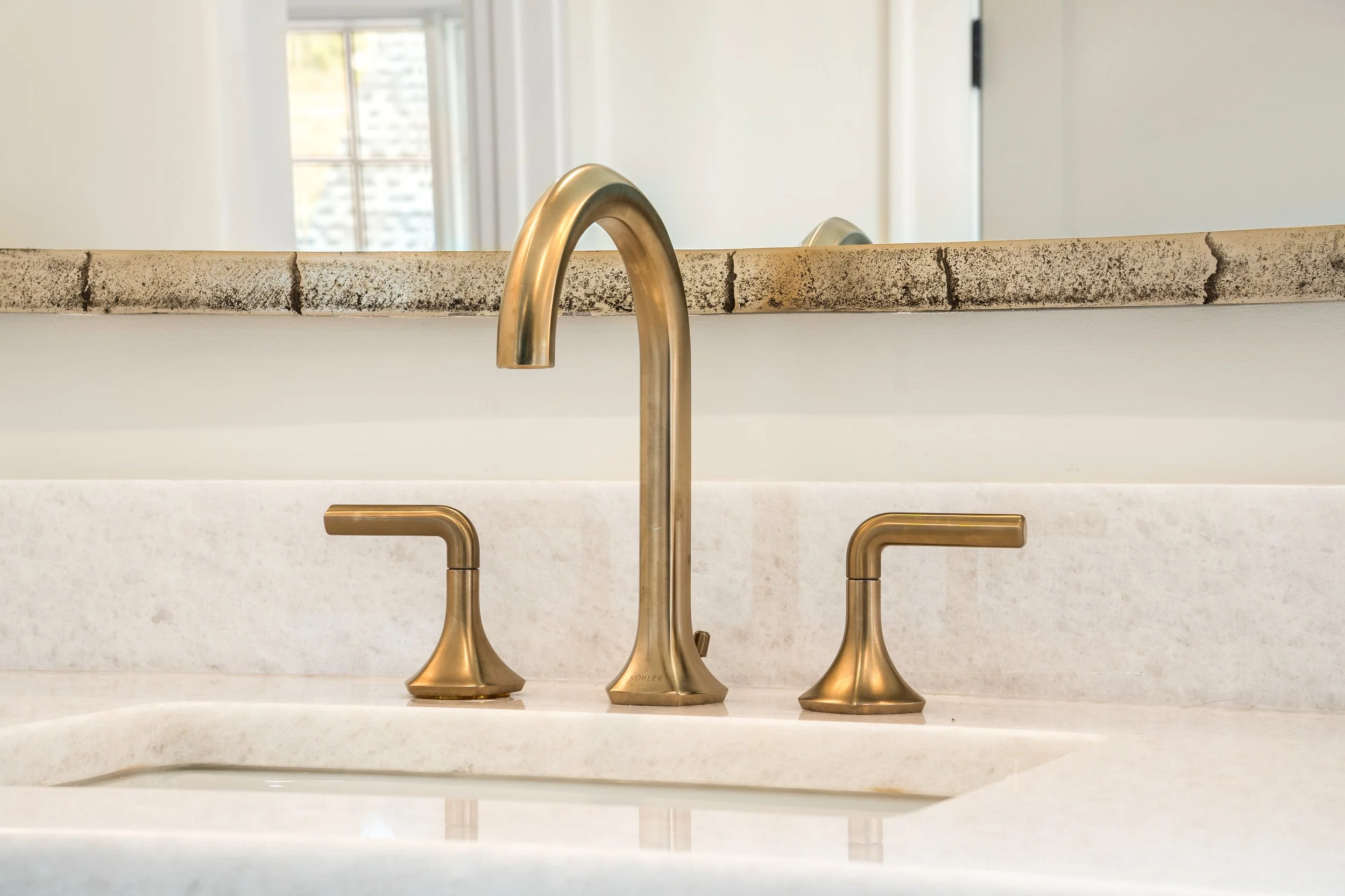 Brass kitchen faucet with curved spout and handles mounted on a white countertop, with a large window in the background.