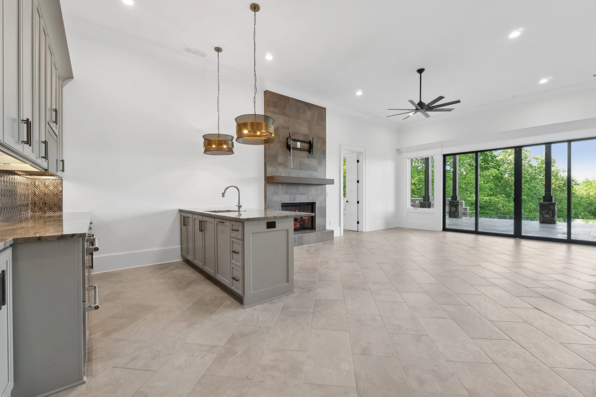 Open-concept living area with tiled floors, a fireplace, large sliding glass doors to a balcony, a kitchen island with a sink, modern cabinets, and pendant lights, with views of green trees outside.