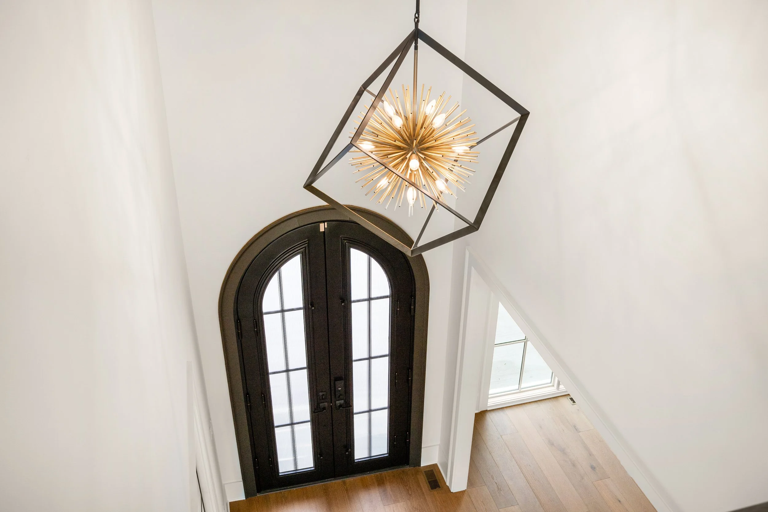 View of a modern interior entryway with a black arched double door window, hardwood flooring, and a geometric chandelier hanging from the ceiling.