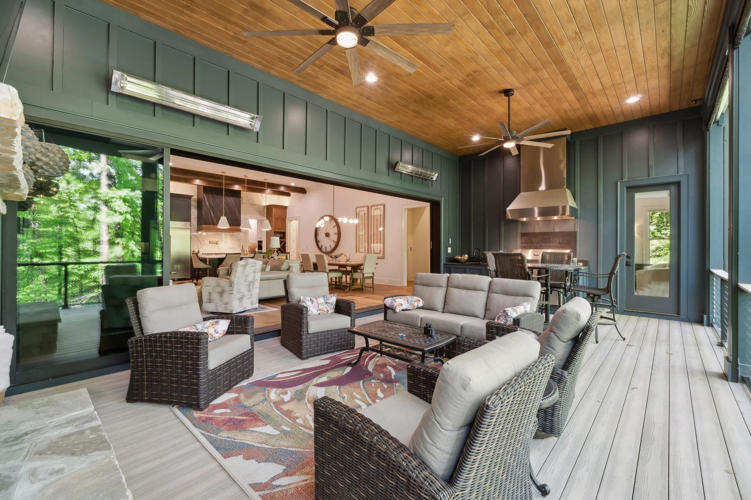 A spacious living area with indoor seating, wicker chairs, a lounge sofa, and a colorful rug. The room features large glass doors that open to a lush green outdoor space, and a kitchen area with pendant lights and a large wall clock is visible in the