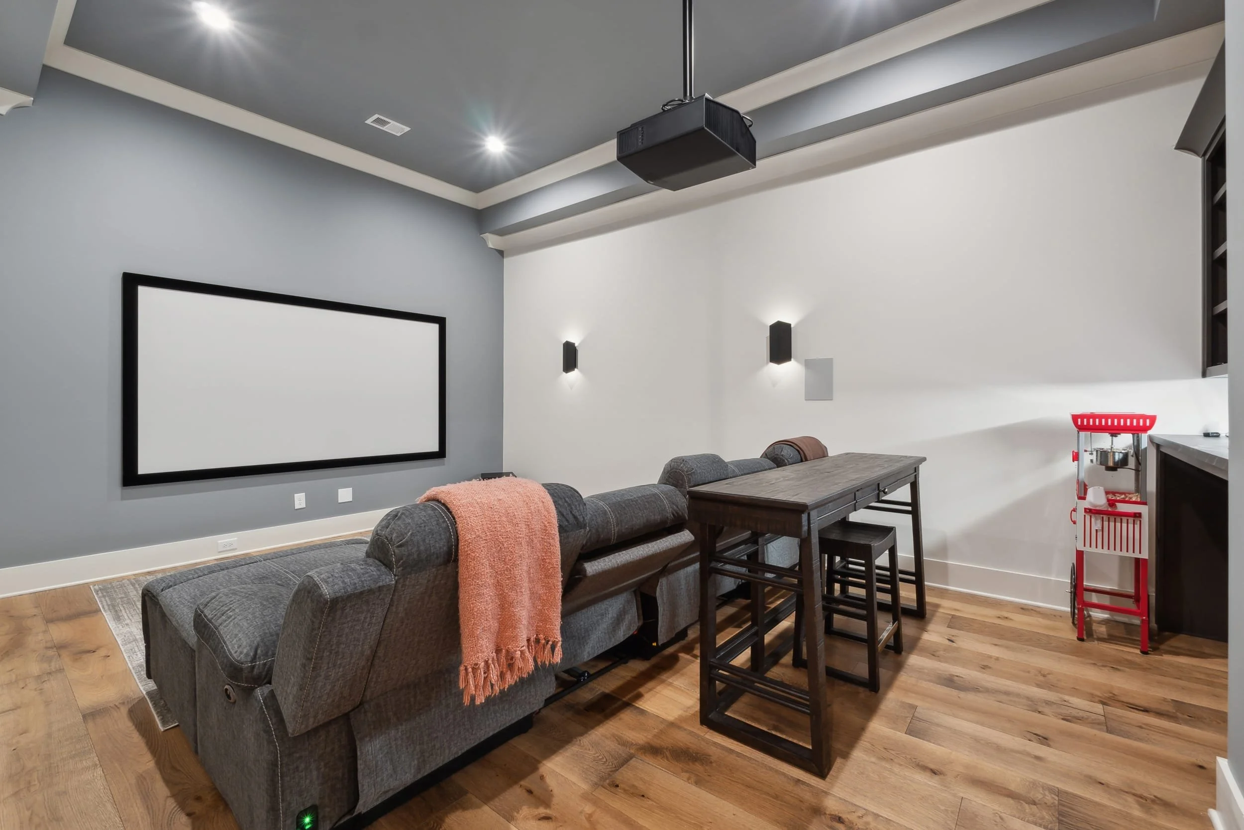 A room with a large flat-screen TV on a white wall, gray sofas with an orange throw, a dining table with chairs, and a red popcorn machine in the corner.
