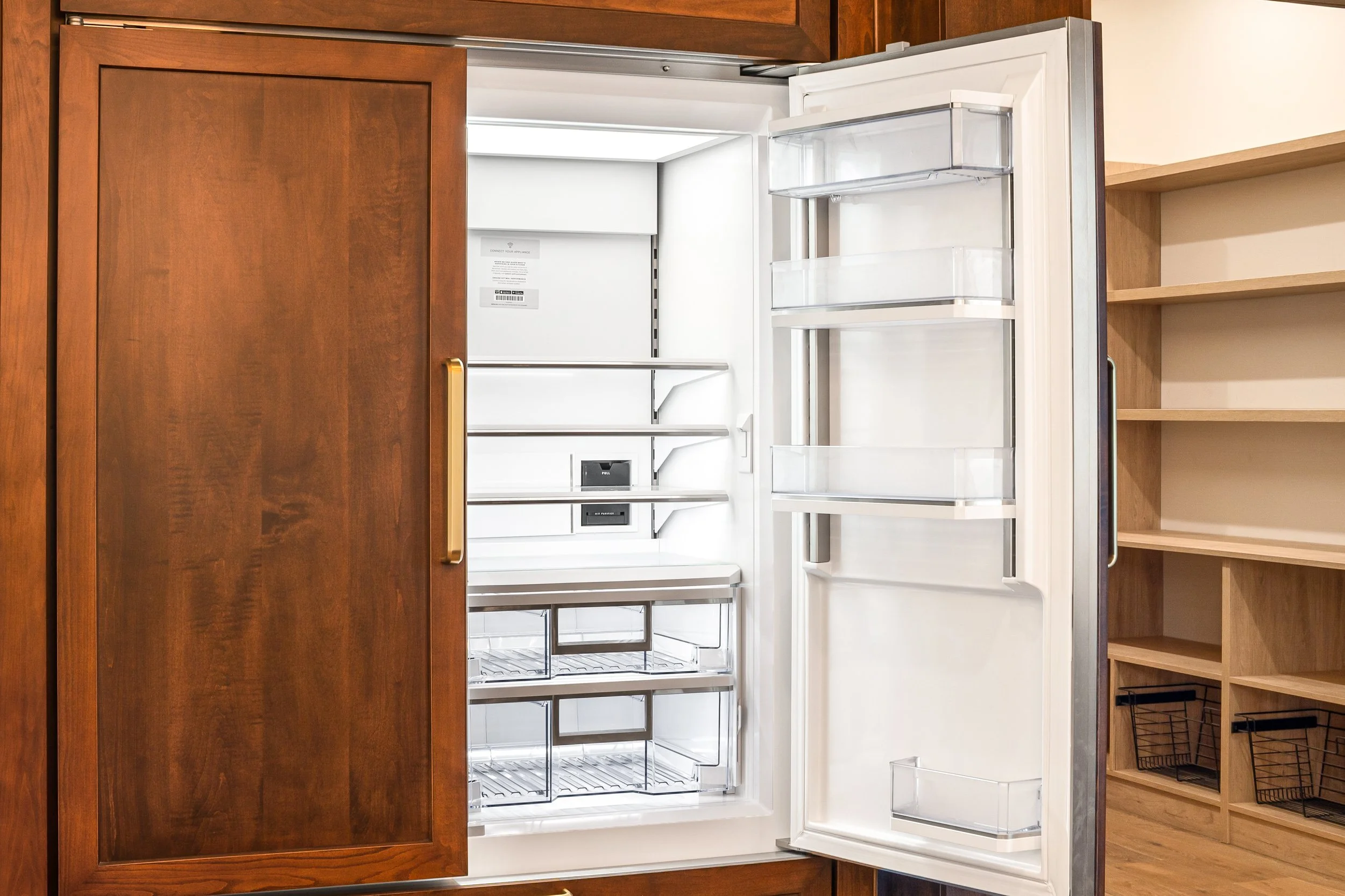 Empty built-in refrigerator with wood cabinet door open, showing white interior with shelves and drawers, in a kitchen with wooden shelves and cabinetry.