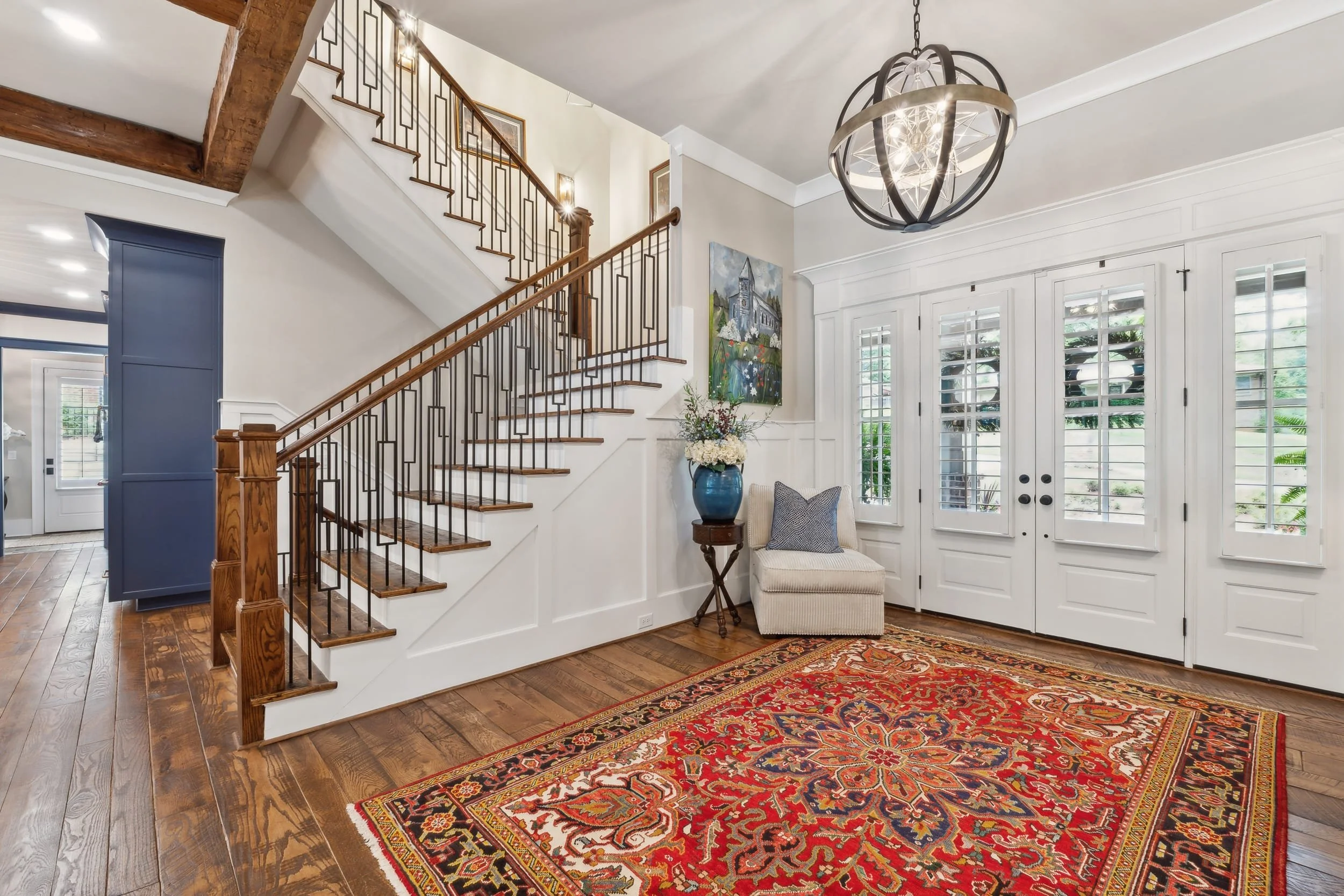 Indoor foyer area showing a staircase with wooden steps and black metal railing, a chandelier hanging from the ceiling, a colorful armchair, a blue vase with flowers, and windows with white shutters.