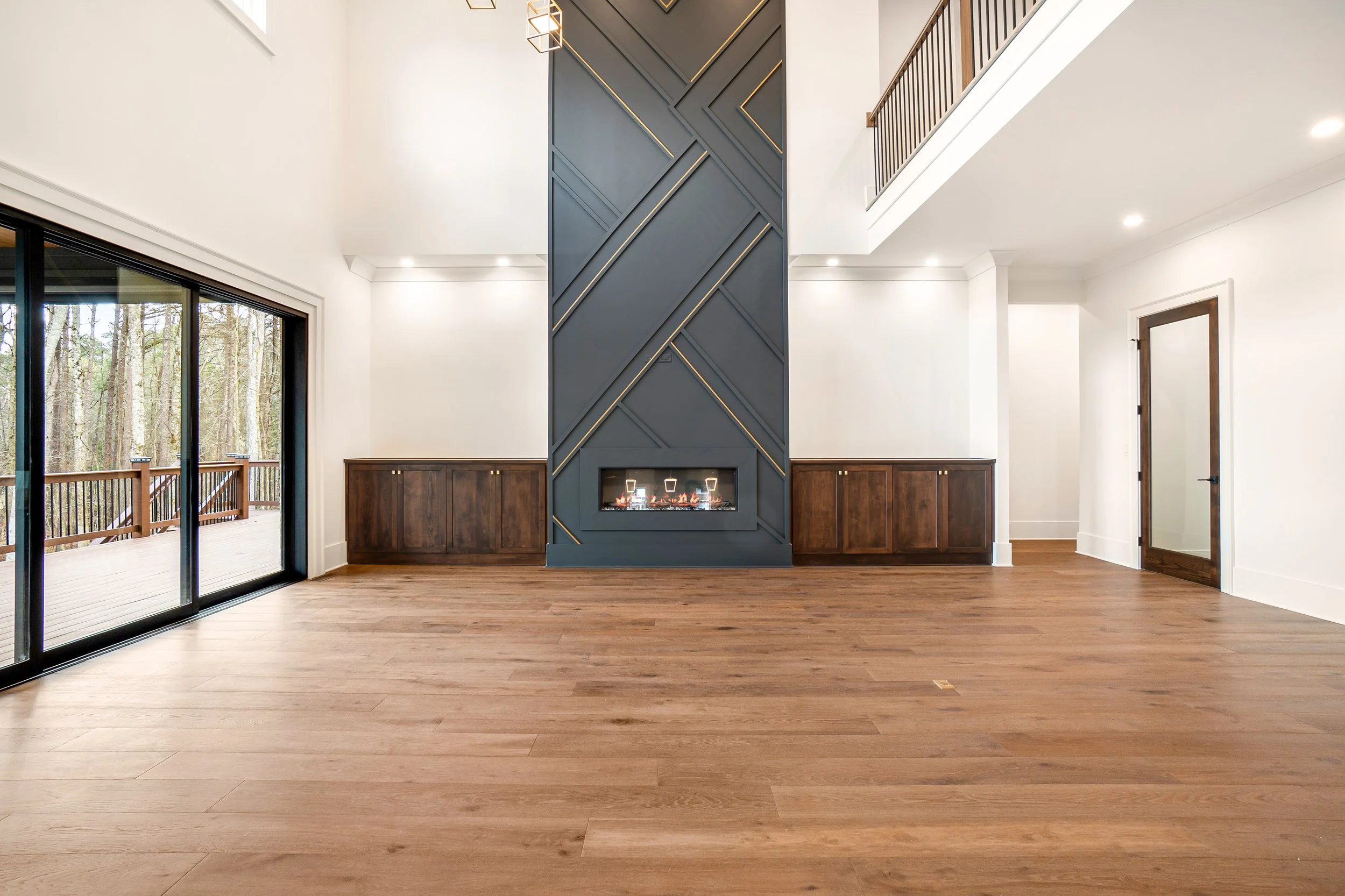 Modern living room with a tall, blue-gray accent wall featuring a geometric design and a built-in fireplace, hardwood flooring, large sliding glass doors leading to an outdoor deck, and a staircase with dark wood railing.