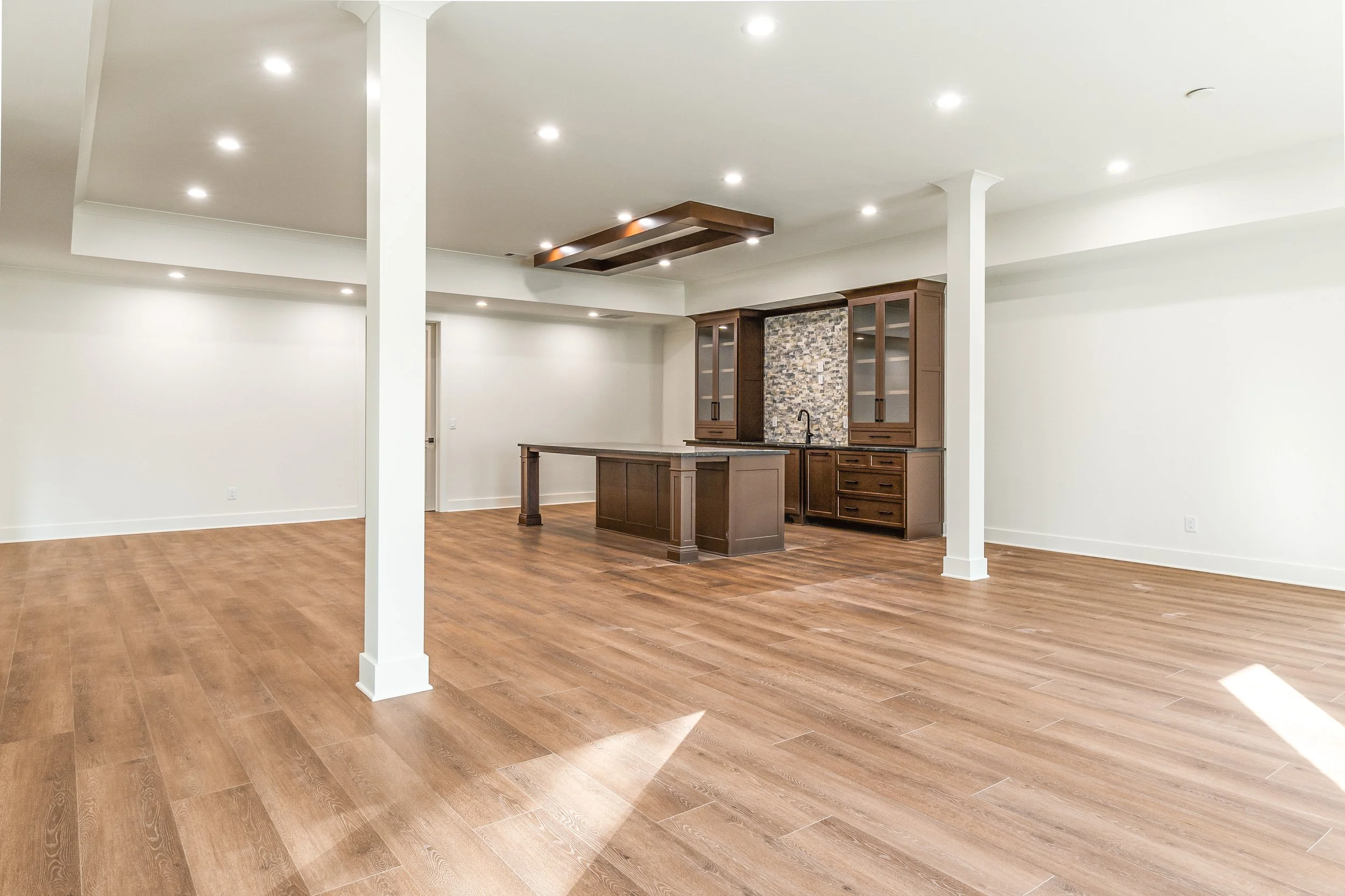 Empty open-concept living space with hardwood floors, white walls, and a kitchen area with dark wood cabinets and a stone backsplash, lit by recessed ceiling lights.