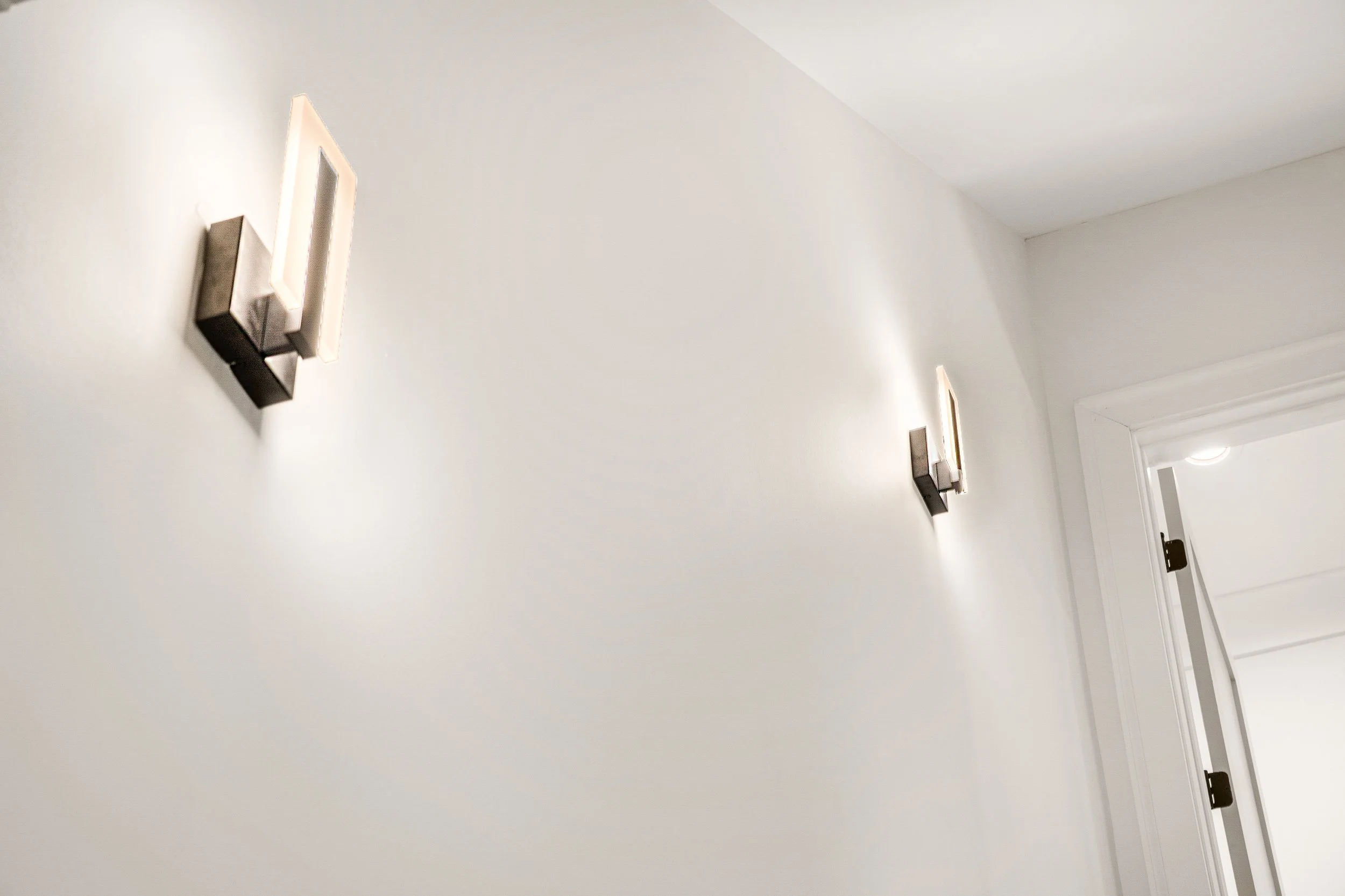 Interior view of a white wall with two modern black wall sconces and a doorframe at the right edge.