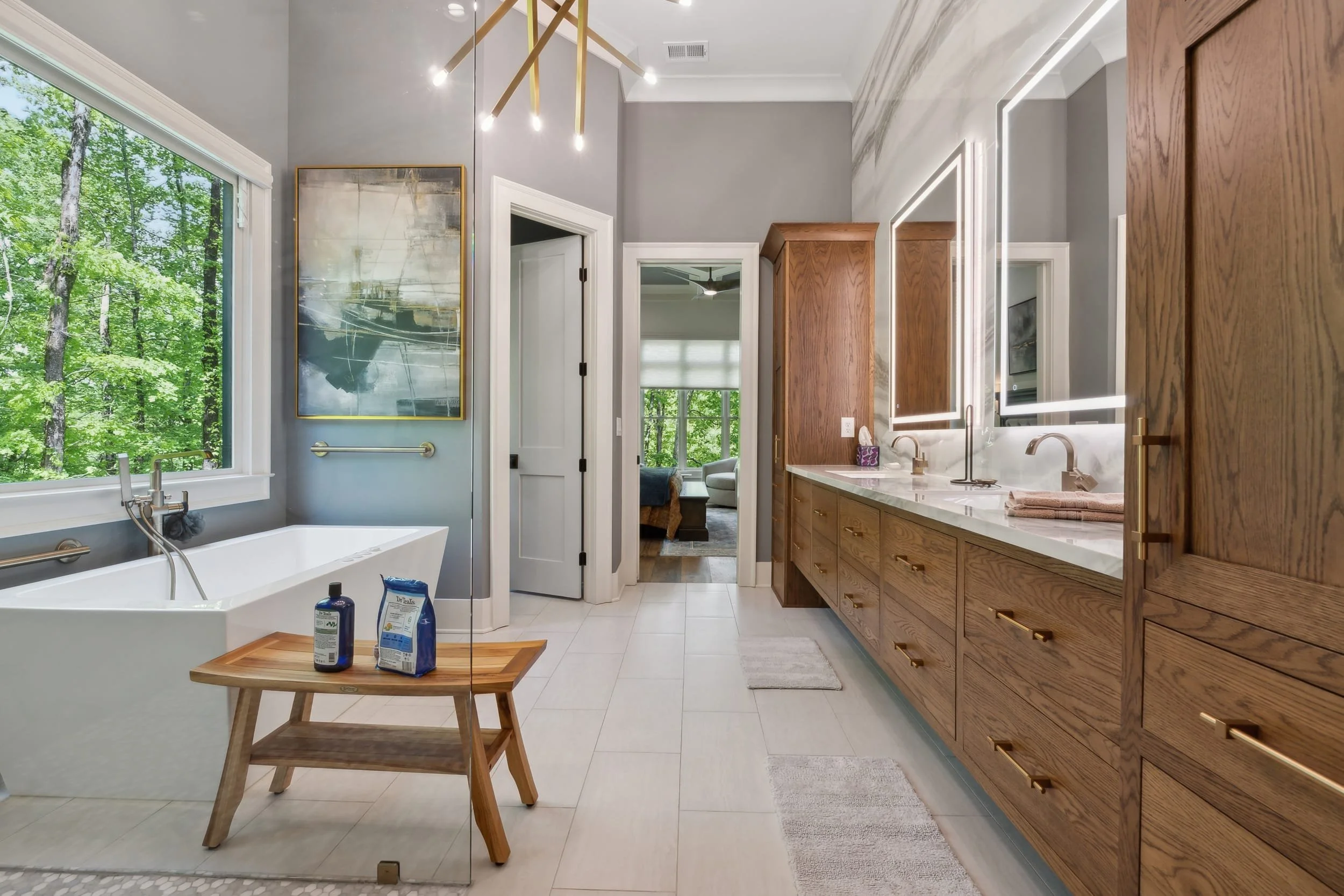 Modern bathroom featuring a large window with a view of green trees, a white bathtub with bath products, a wooden bench, and a double vanity with lit mirrors and wooden cabinets.