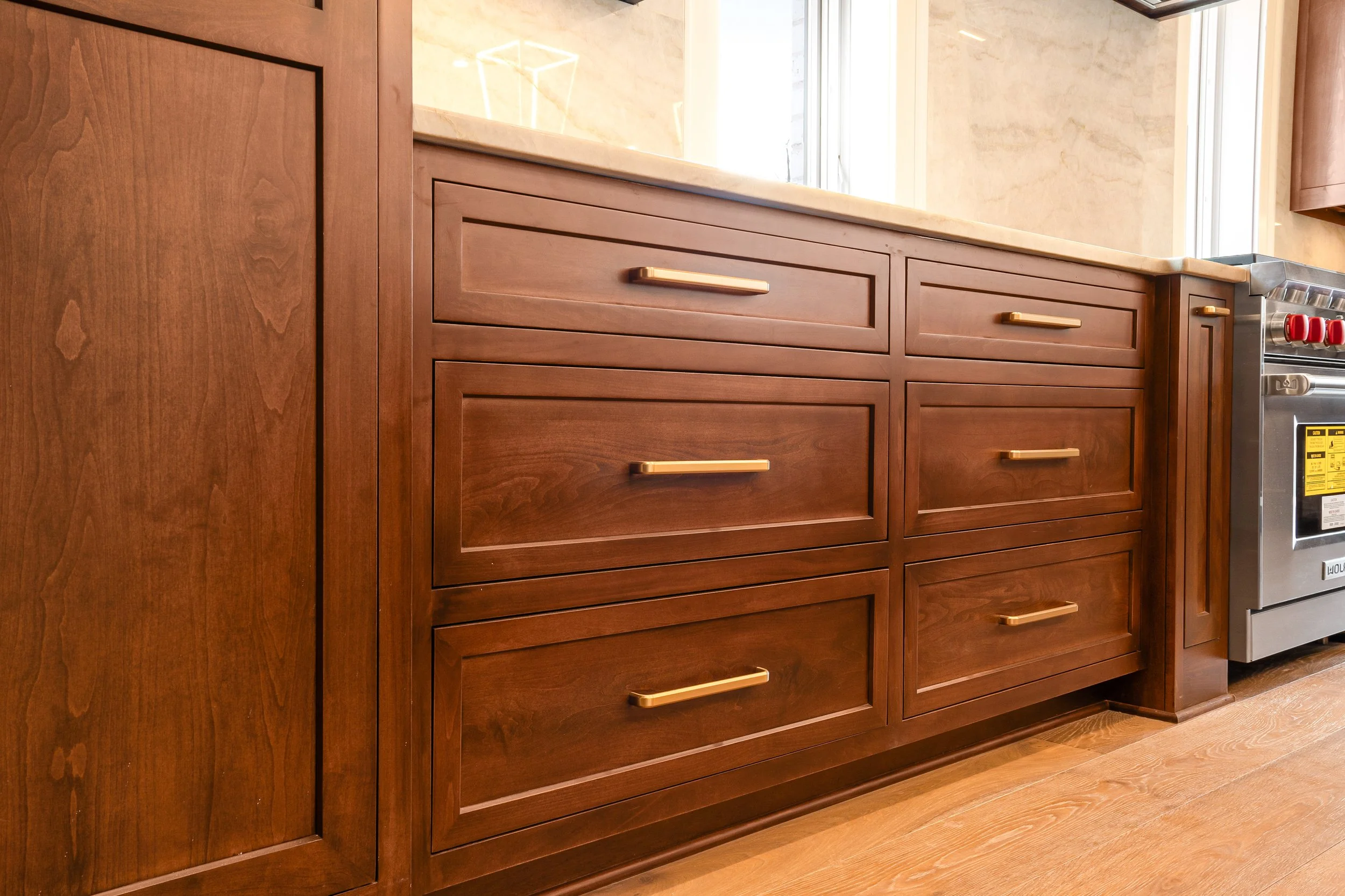 Close-up of a kitchen cabinet with wooden finish and gold-colored handles, part of a kitchen setup including a marble countertop, with a window above and a portion of a metallic appliance to the right.