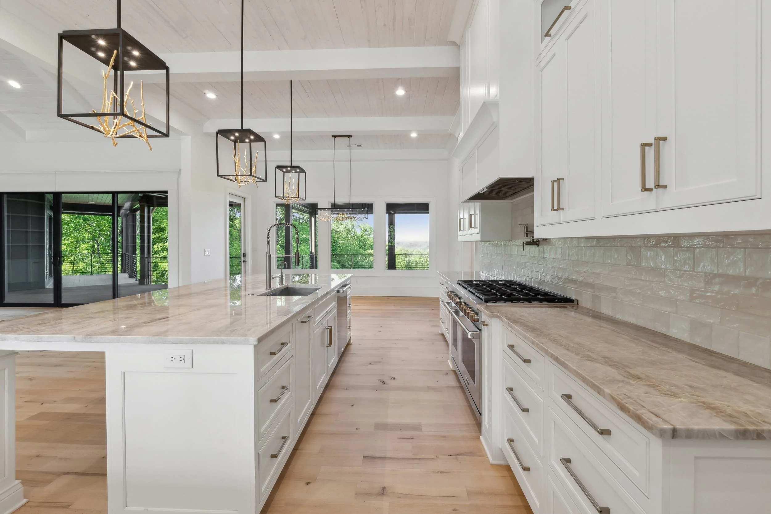 Modern kitchen with white cabinets, marble countertops, pendant lights with branch motifs, large windows showing greenery outside, and light wooden flooring.