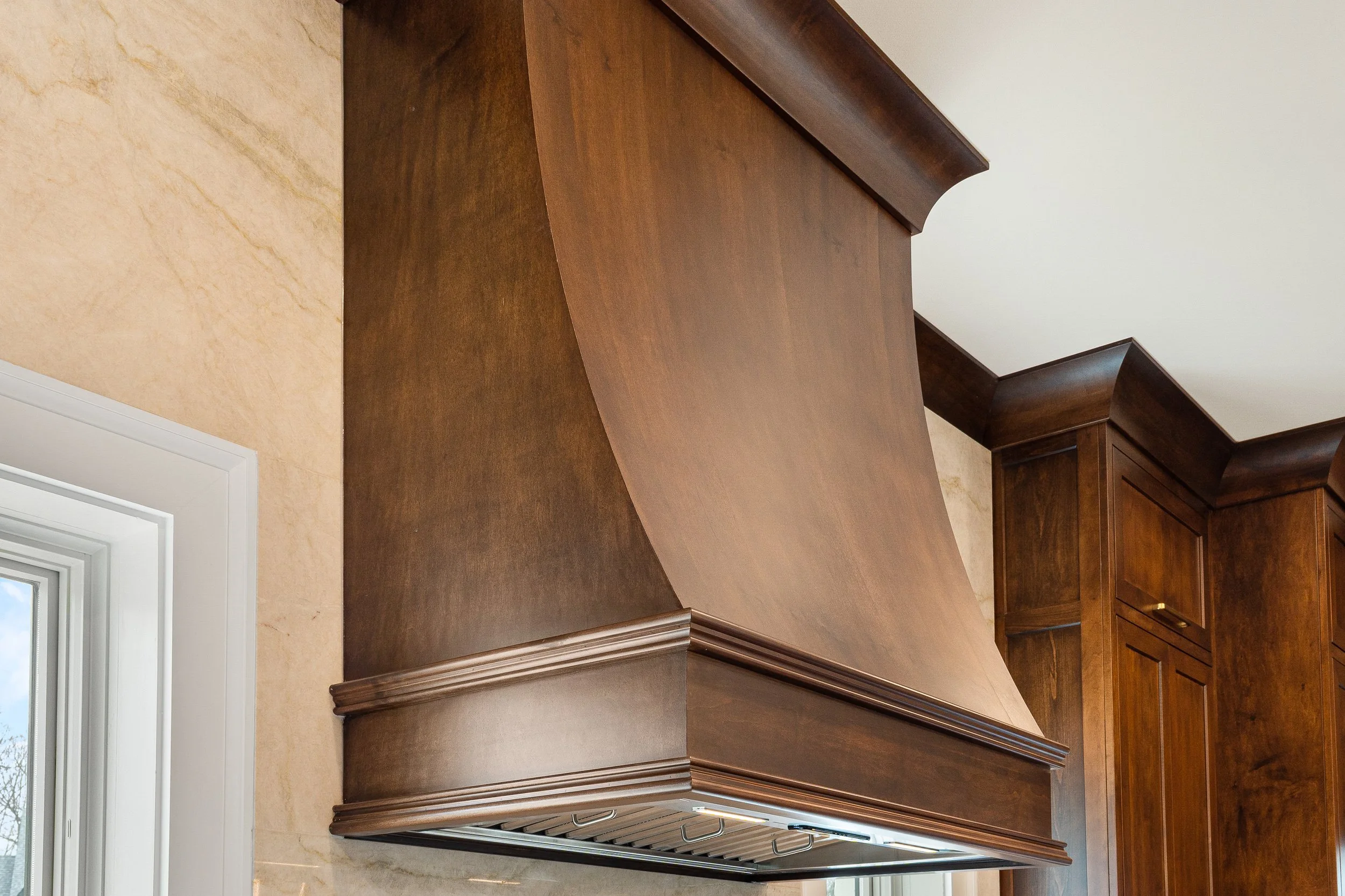 View of wooden kitchen cabinets with a crown molding top edge, seen from below, with a beige marble wall and a white window frame to the left.