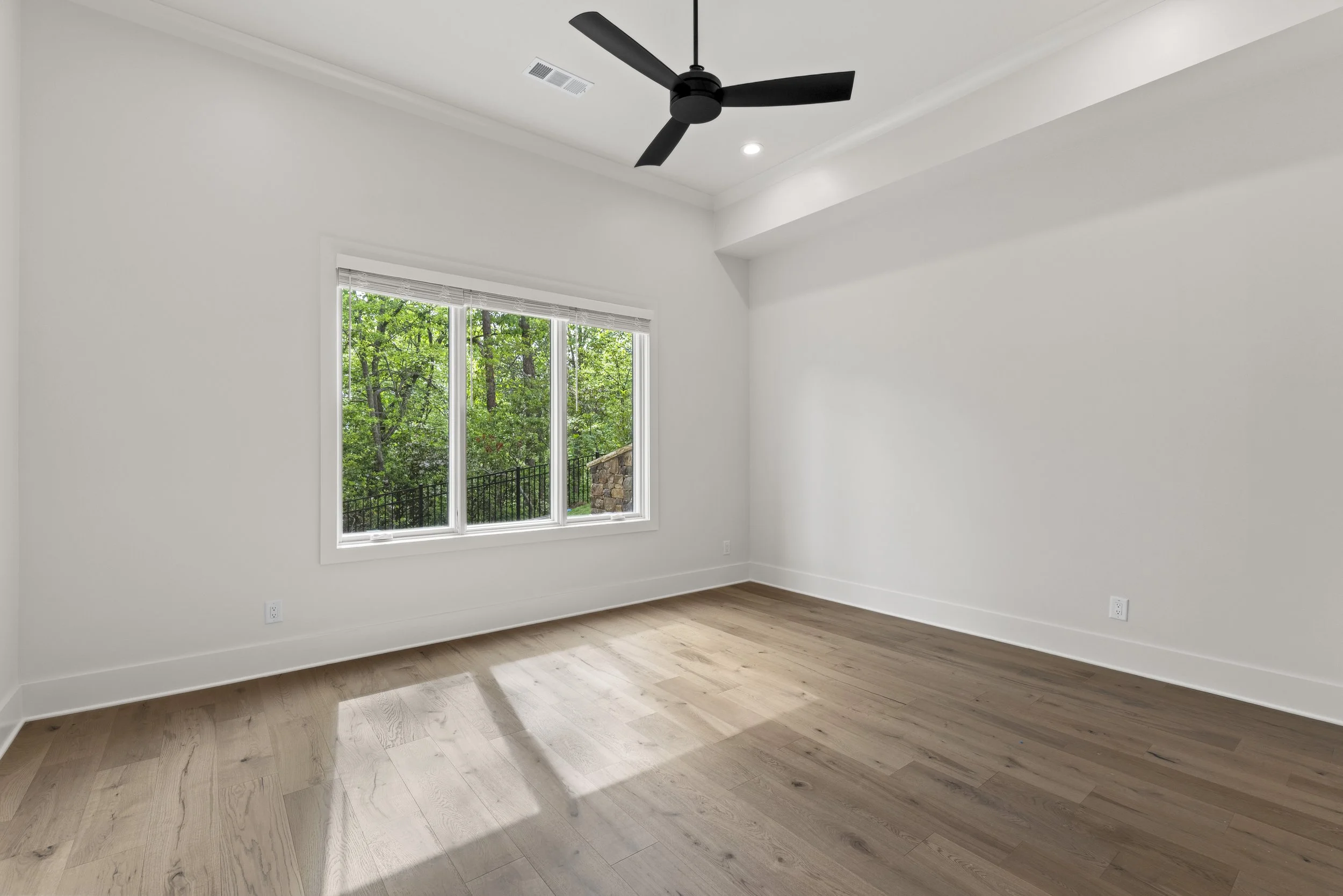 Empty room with white walls, wooden floor, large window with views of green trees, ceiling fan, and recessed lighting.