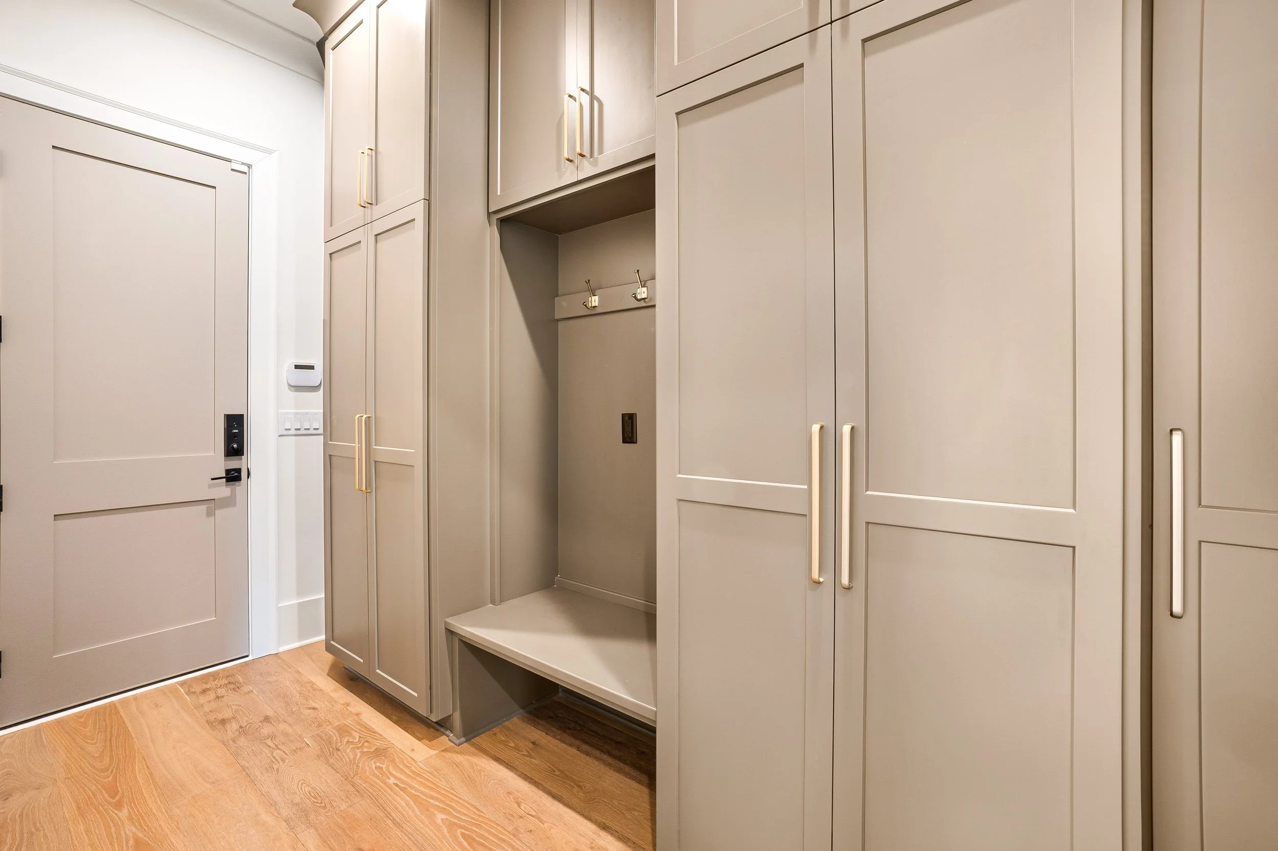 A hallway with beige cabinetry, gold handles, and a bench area with hooks, on a hardwood floor.