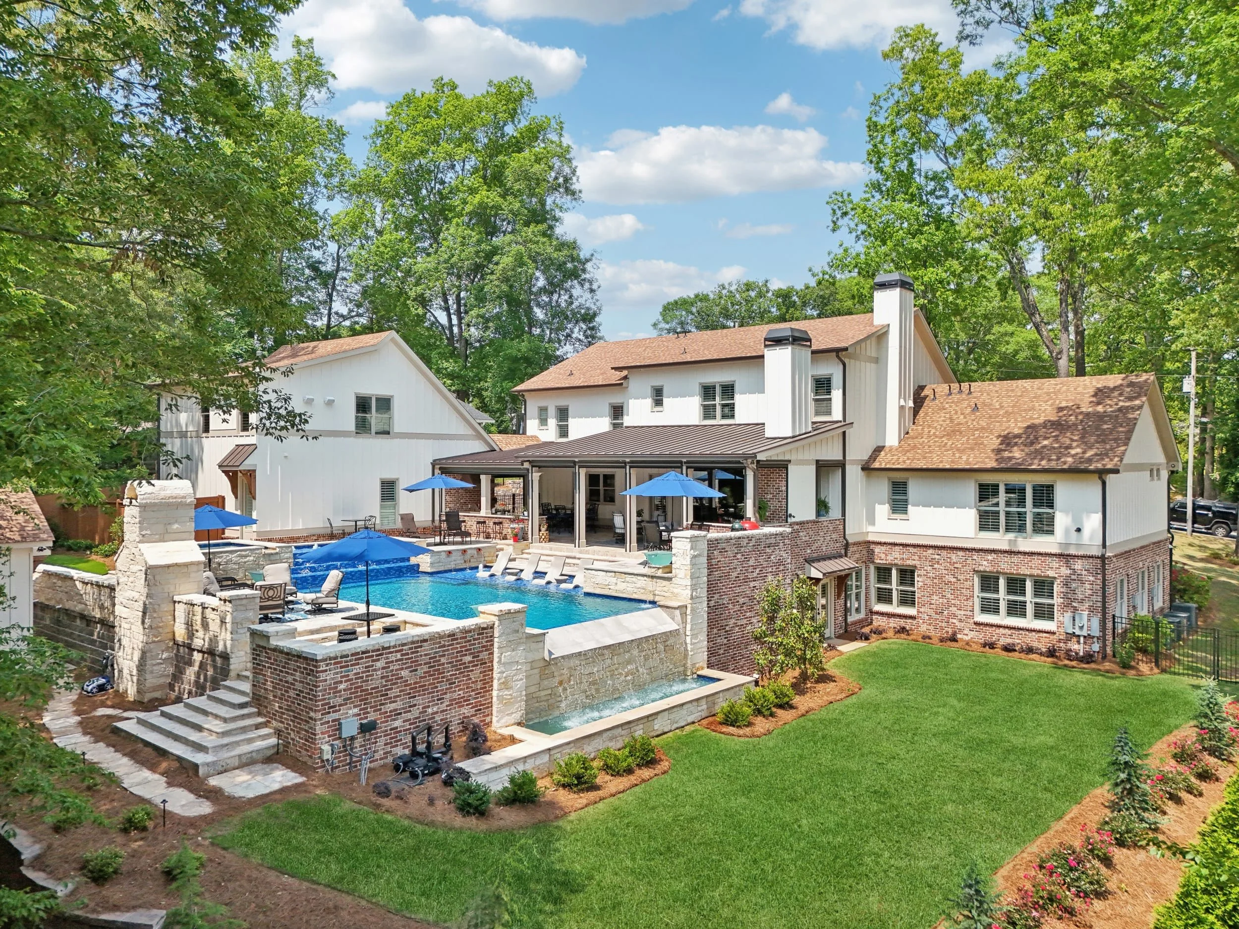 A backyard featuring a swimming pool surrounded by a brick wall, patio furniture with umbrellas, and a two-story house with white siding and a brown roof, with trees and a blue sky in the background.