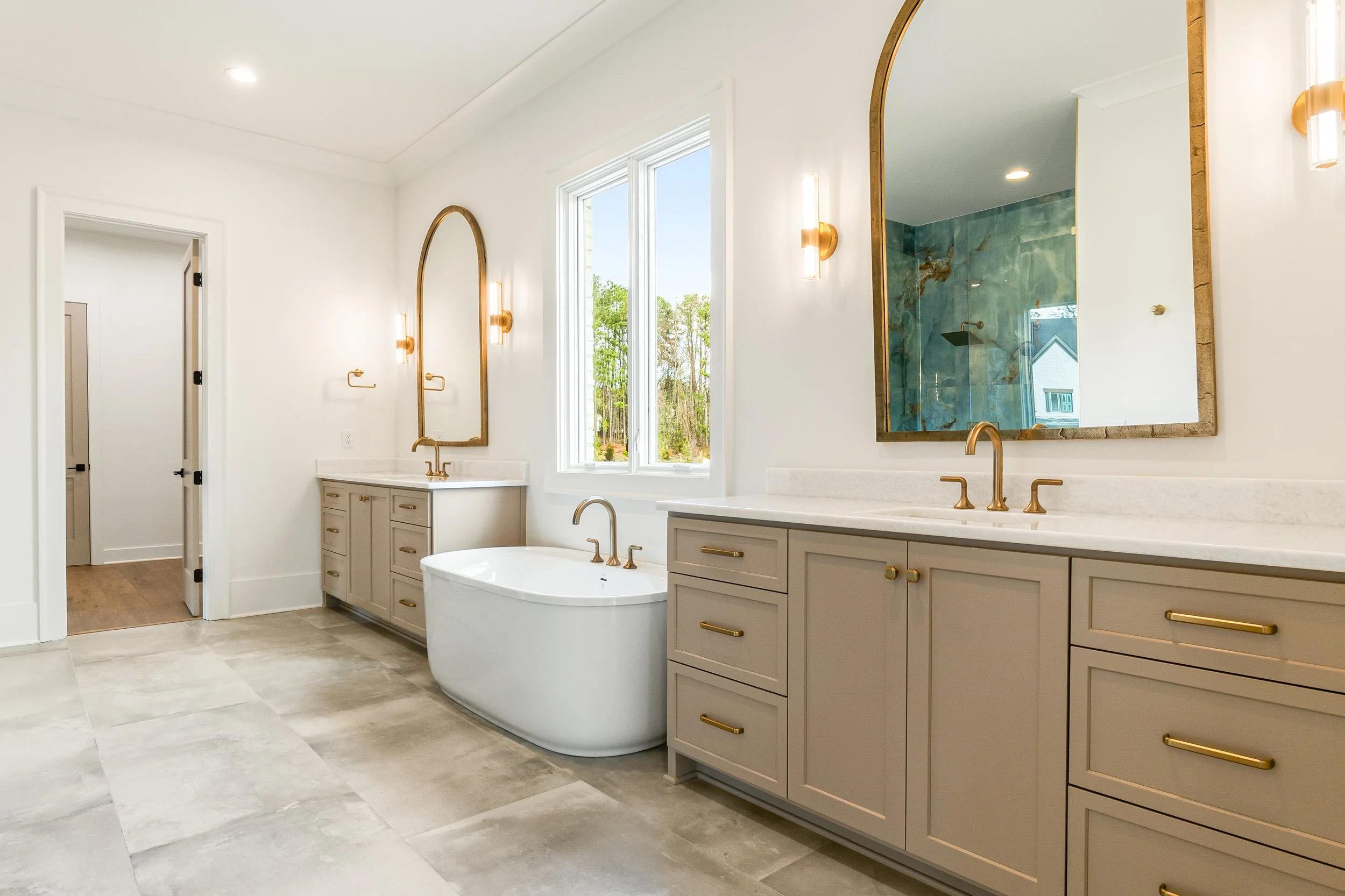 Modern bathroom with double vanity, gold fixtures, large mirrors, bathtub, window with outdoor view, and walk-in shower with blue marble tiles.