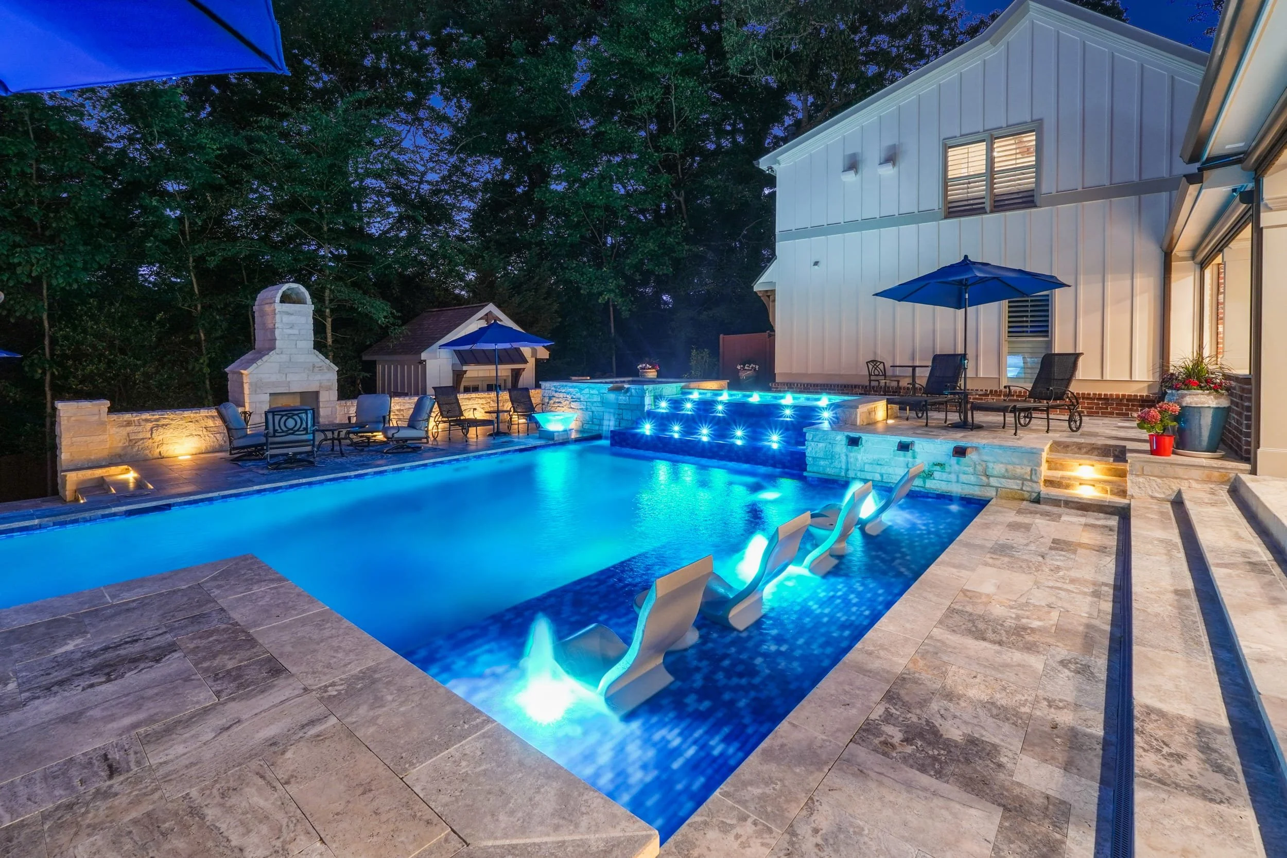 A backyard swimming pool area lit at dusk with blue lighting, surrounded by patio furniture, umbrellas, and a white house with large windows. Trees are visible in the background.