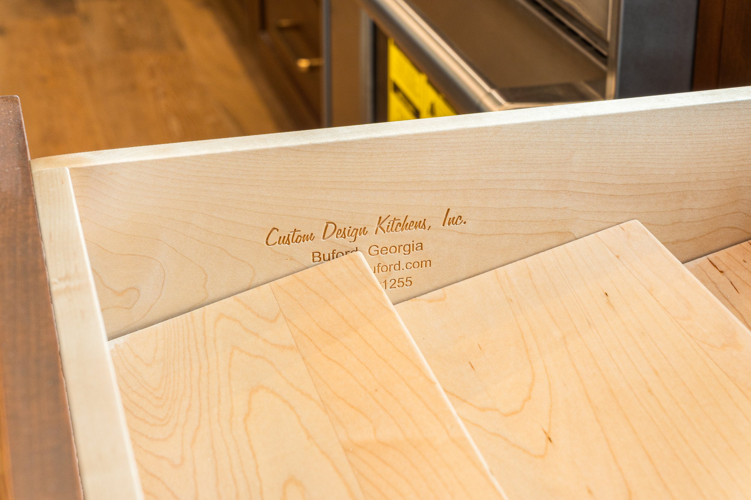 Close-up of a light-colored wooden drawer with engraved text, partially open, revealing a cabinet or appliance with a yellow label in a kitchen setting.