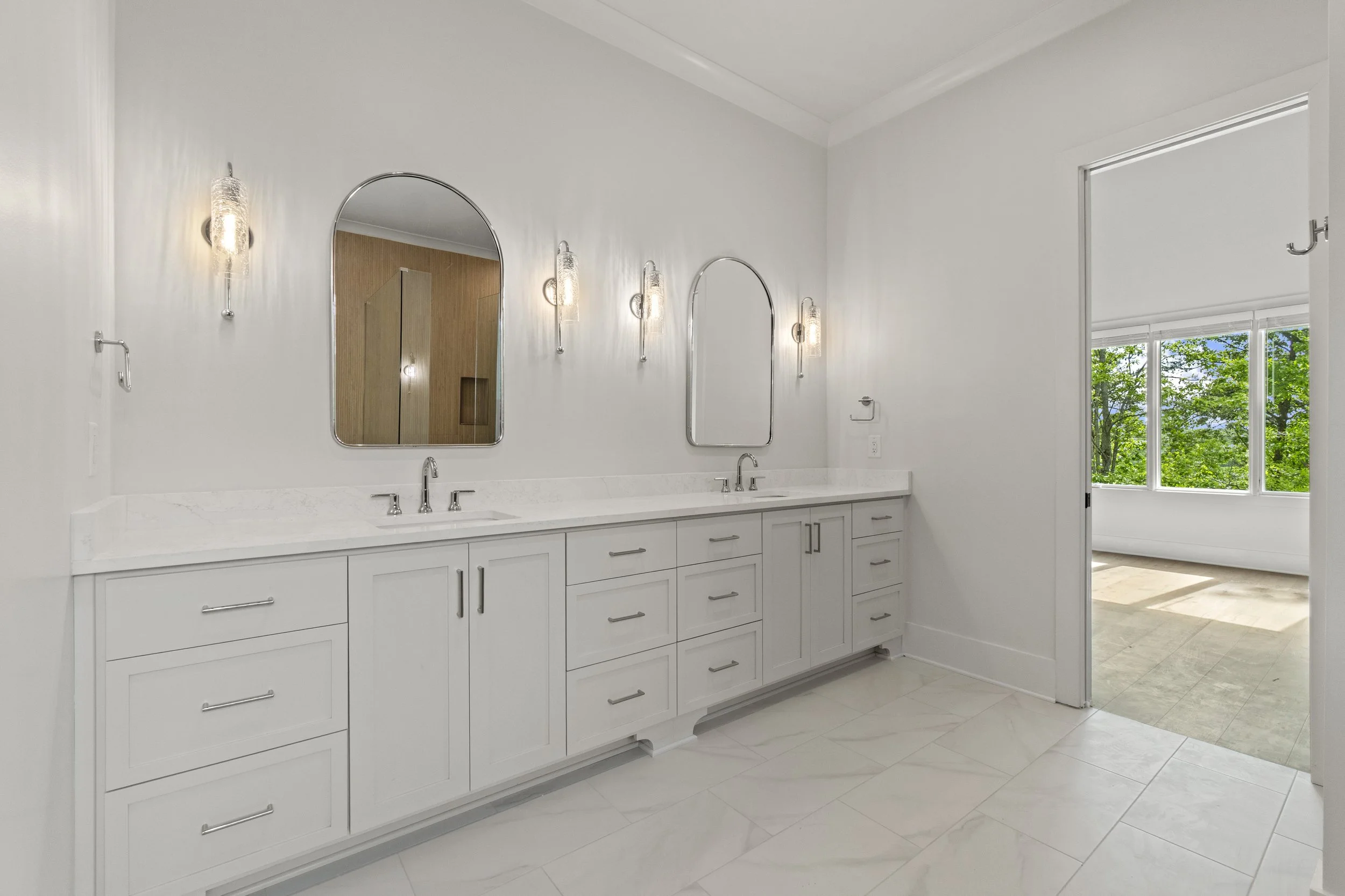 A modern bathroom with white cabinetry and countertops, three mirrors above the sinks, and wall-mounted light fixtures. A window with trees outside is visible on the right.