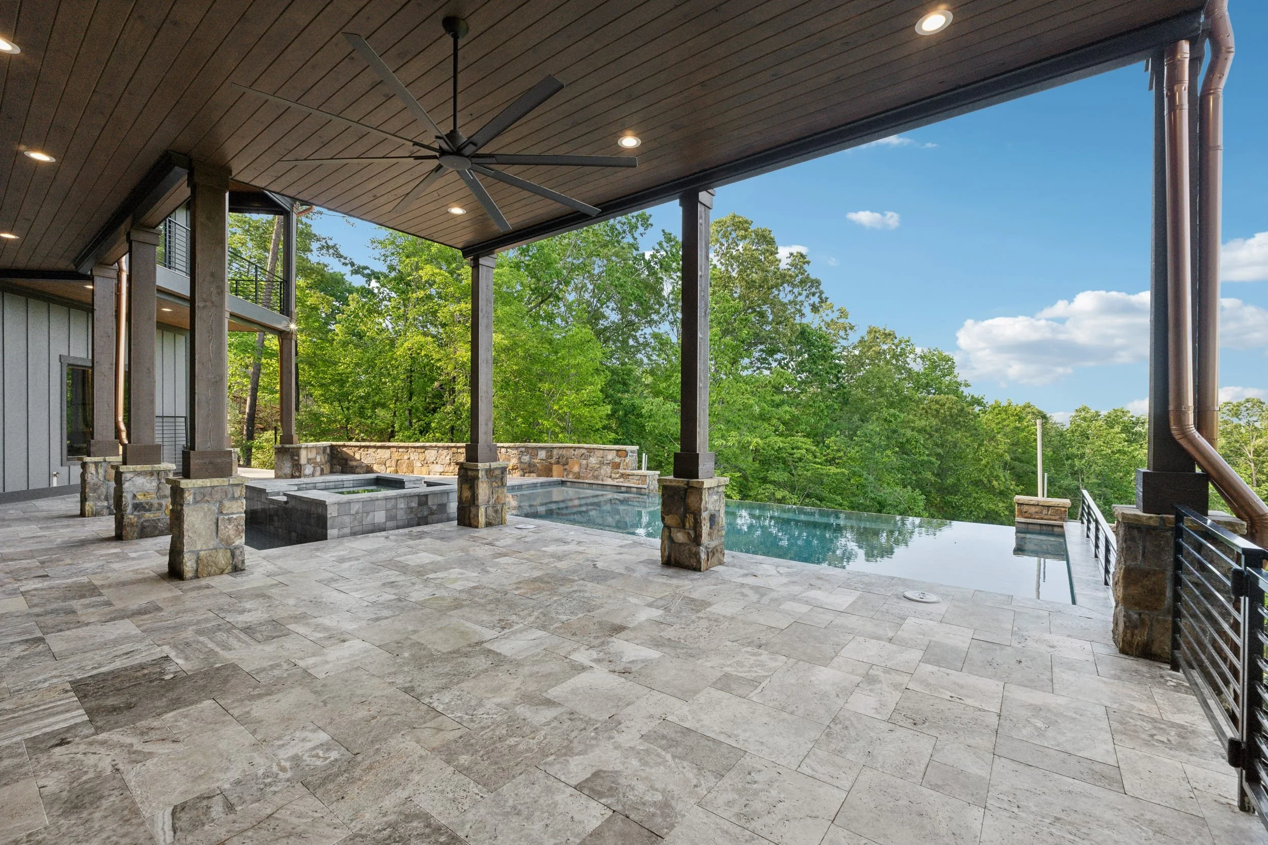 Covered outdoor patio with stone flooring, ceiling fan, recessed lighting, and a view of a lush green forest and blue sky.