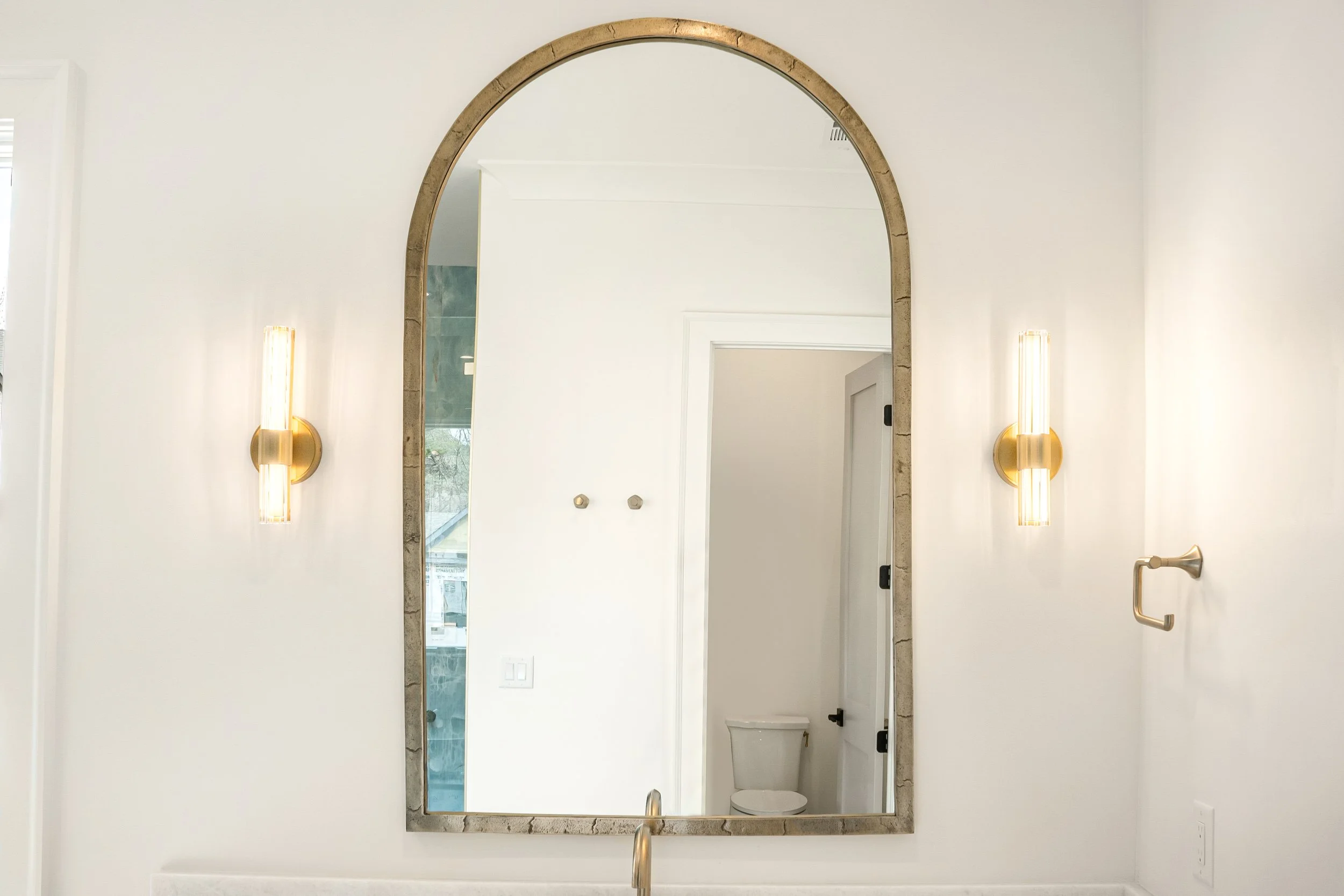 Modern bathroom with an arched mirror, wall-mounted lights, and a glimpse of a toilet through an open door.