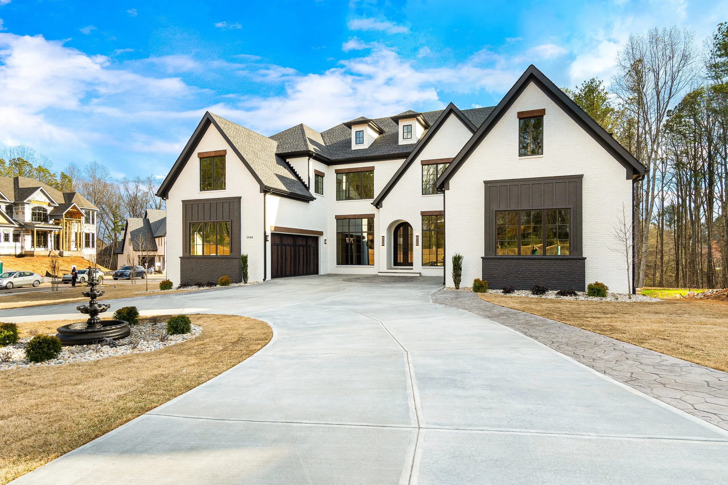 Modern white house with dark trim and a two-car garage, surrounded by a driveway, small trees, and a landscaped yard, under a partly cloudy sky.