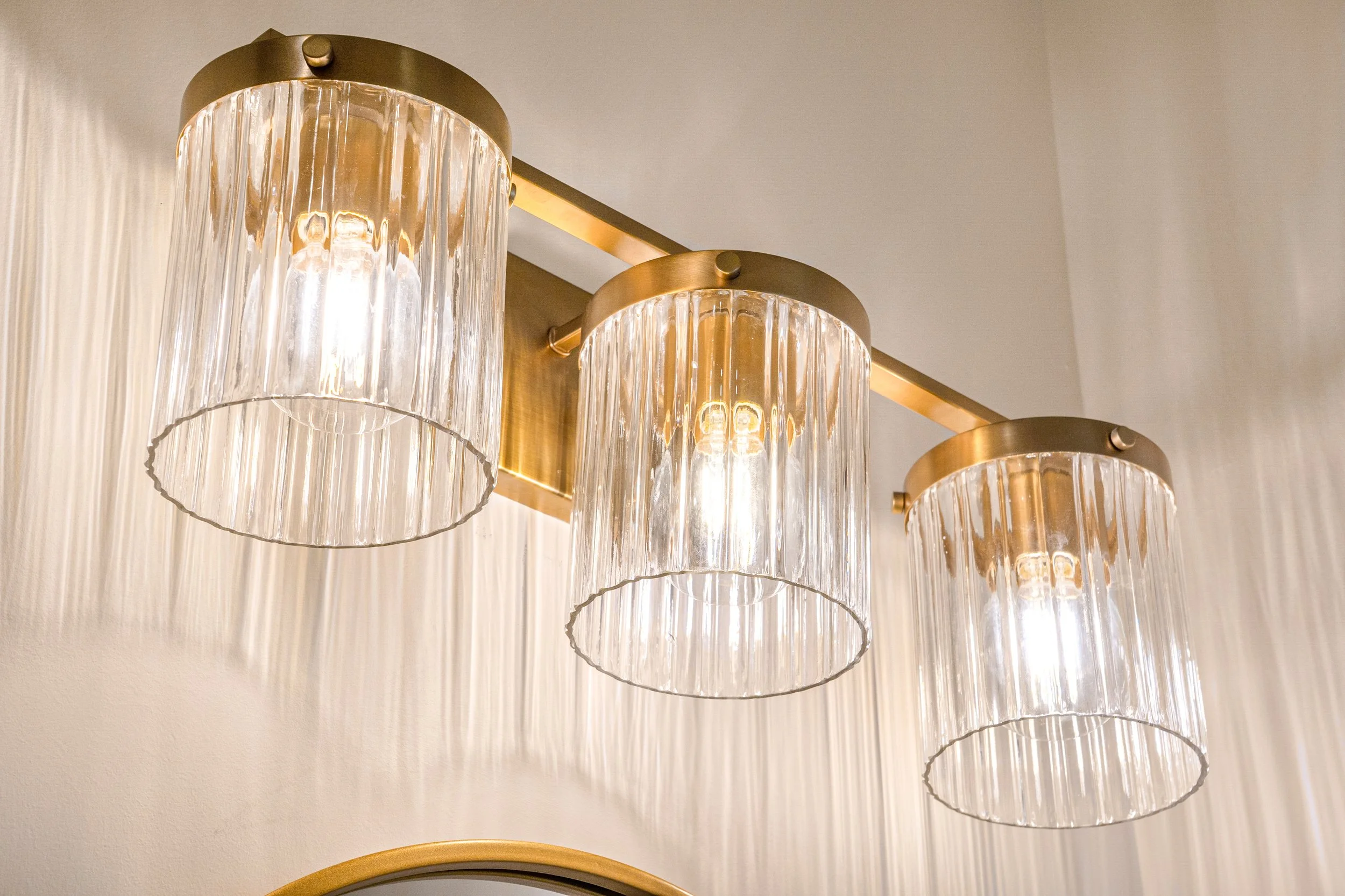 Close-up of three vintage-style pendant lights with glass or crystal shades and brass fixtures hanging from a brass rod against a cream-colored wall.