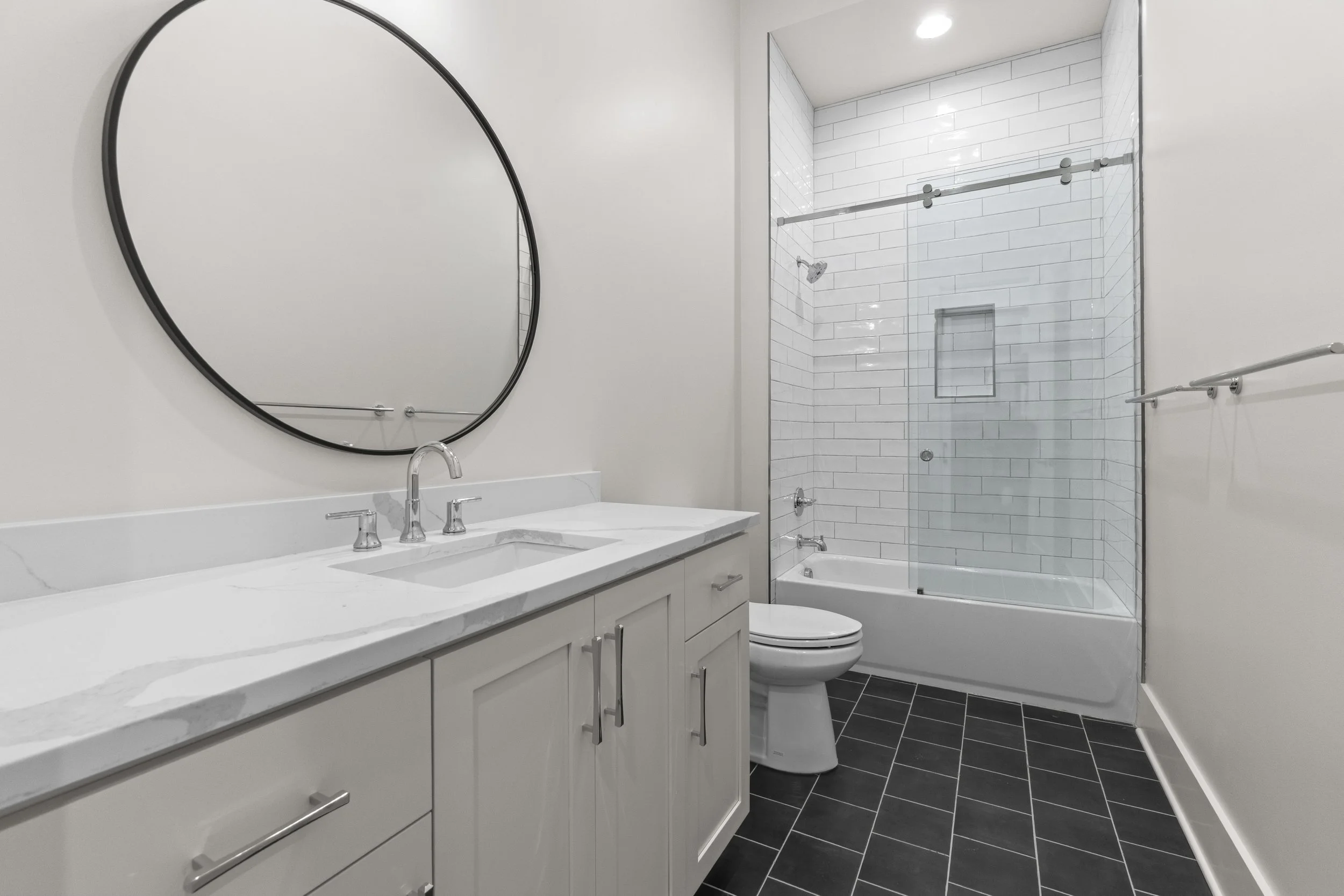 Modern bathroom featuring a white vanity with a marble countertop, a large oval mirror, a toilet, and a glass-enclosed shower with white subway tile walls and black floor tiles.