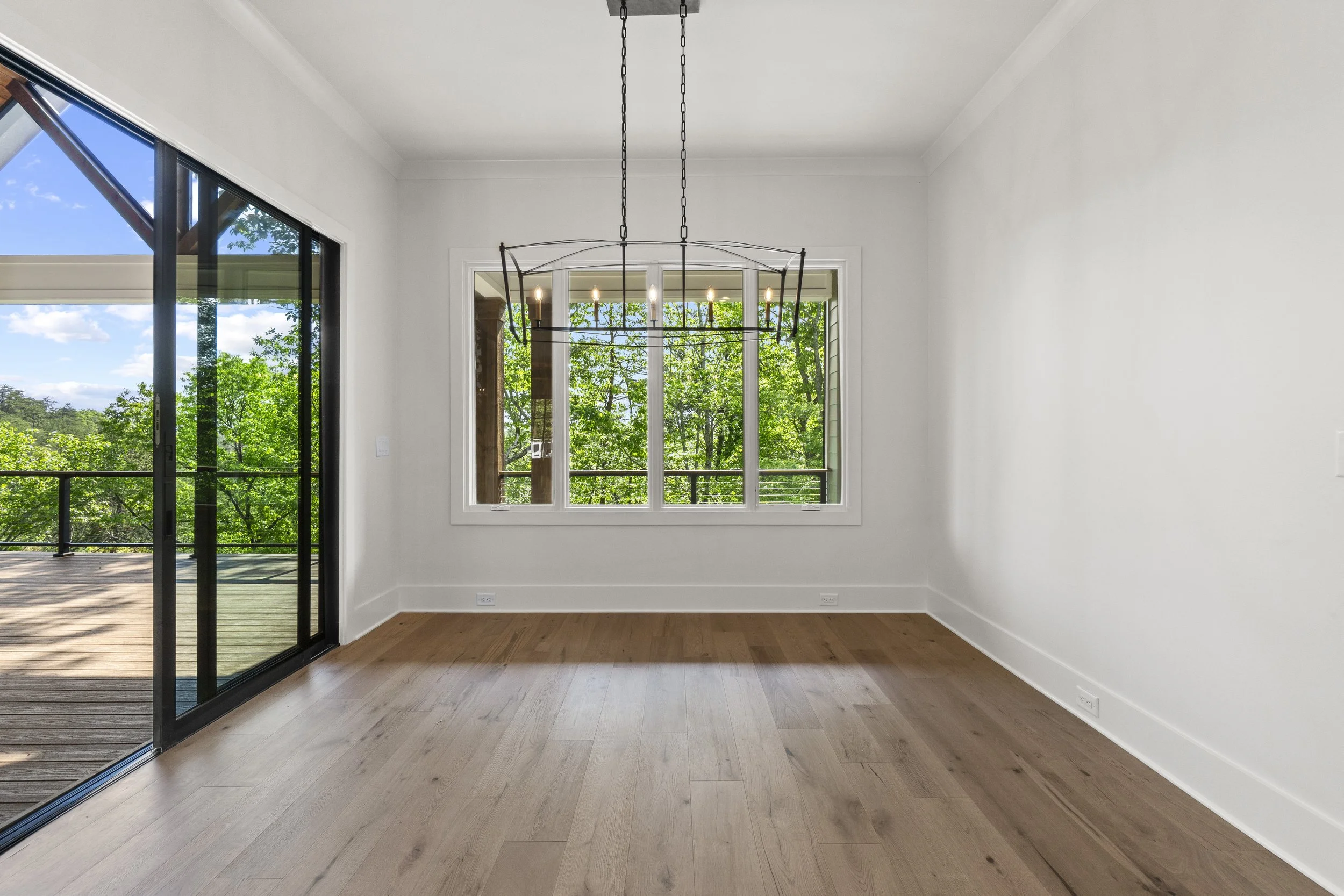 Empty room with hardwood flooring, white walls, large window with a view of green trees, and an elegant black chandelier hanging from the ceiling. Sliding glass door leading to an outdoor deck.