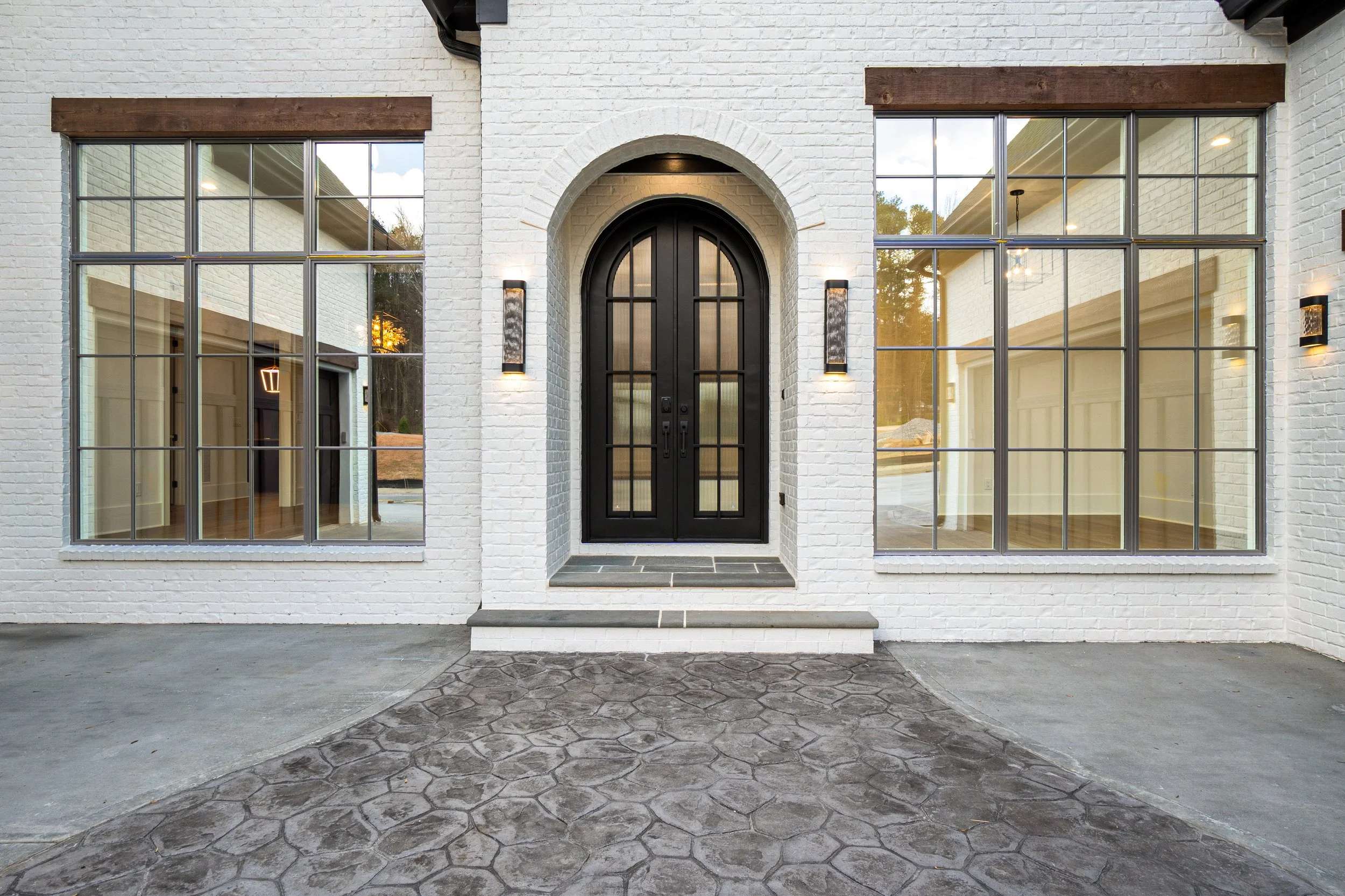 Front view of a modern house with white brick walls, large glass windows, and a black arched front door with two wall-mounted lights on either side, and a small stone project step and paved front yard.