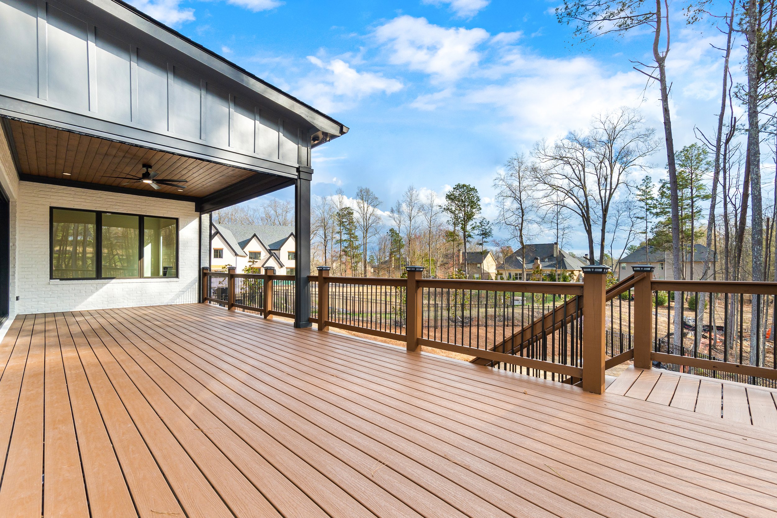 A spacious wooden deck attached to a house with a large window, overlooking a neighborhood with trees and houses under a partly cloudy sky.