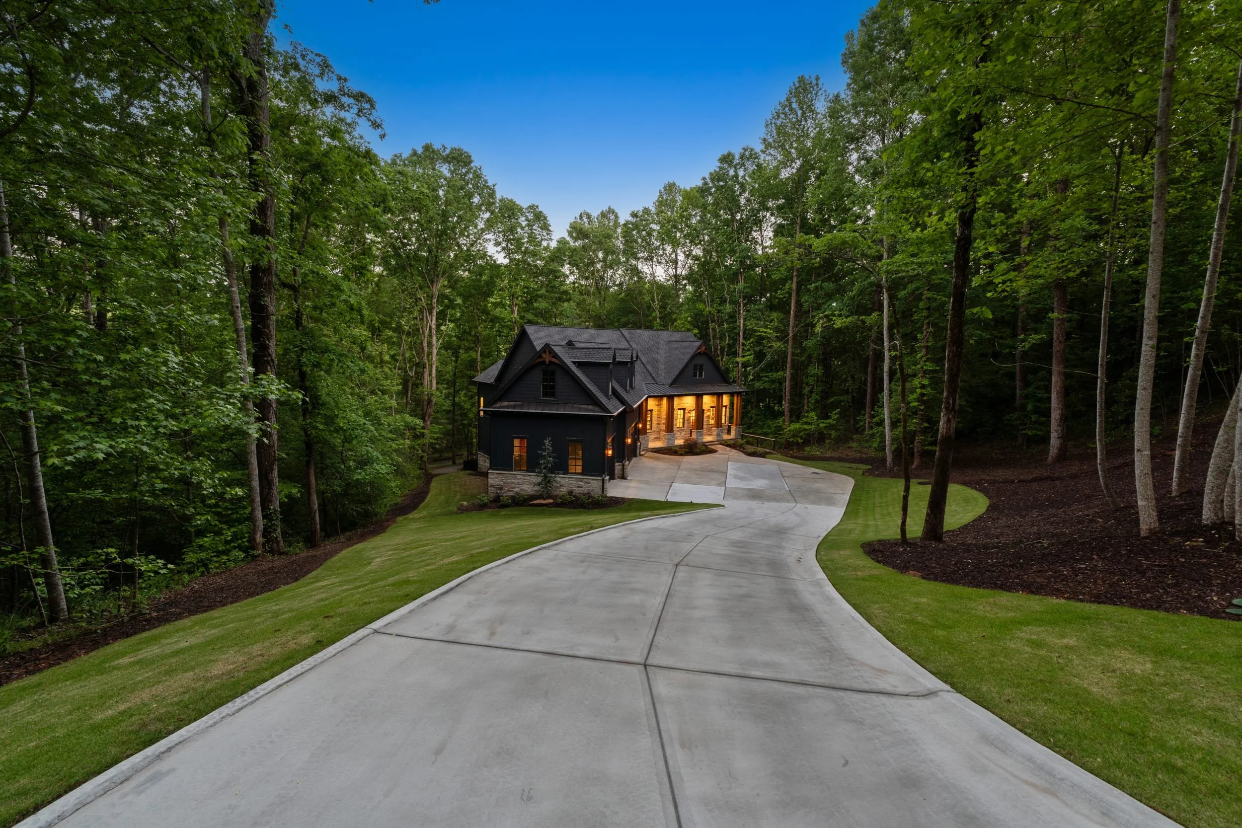 A house nestled in a dense wooded area surrounded by tall trees and a well-maintained lawn, with a concrete driveway leading to the house, illuminated from within during twilight.