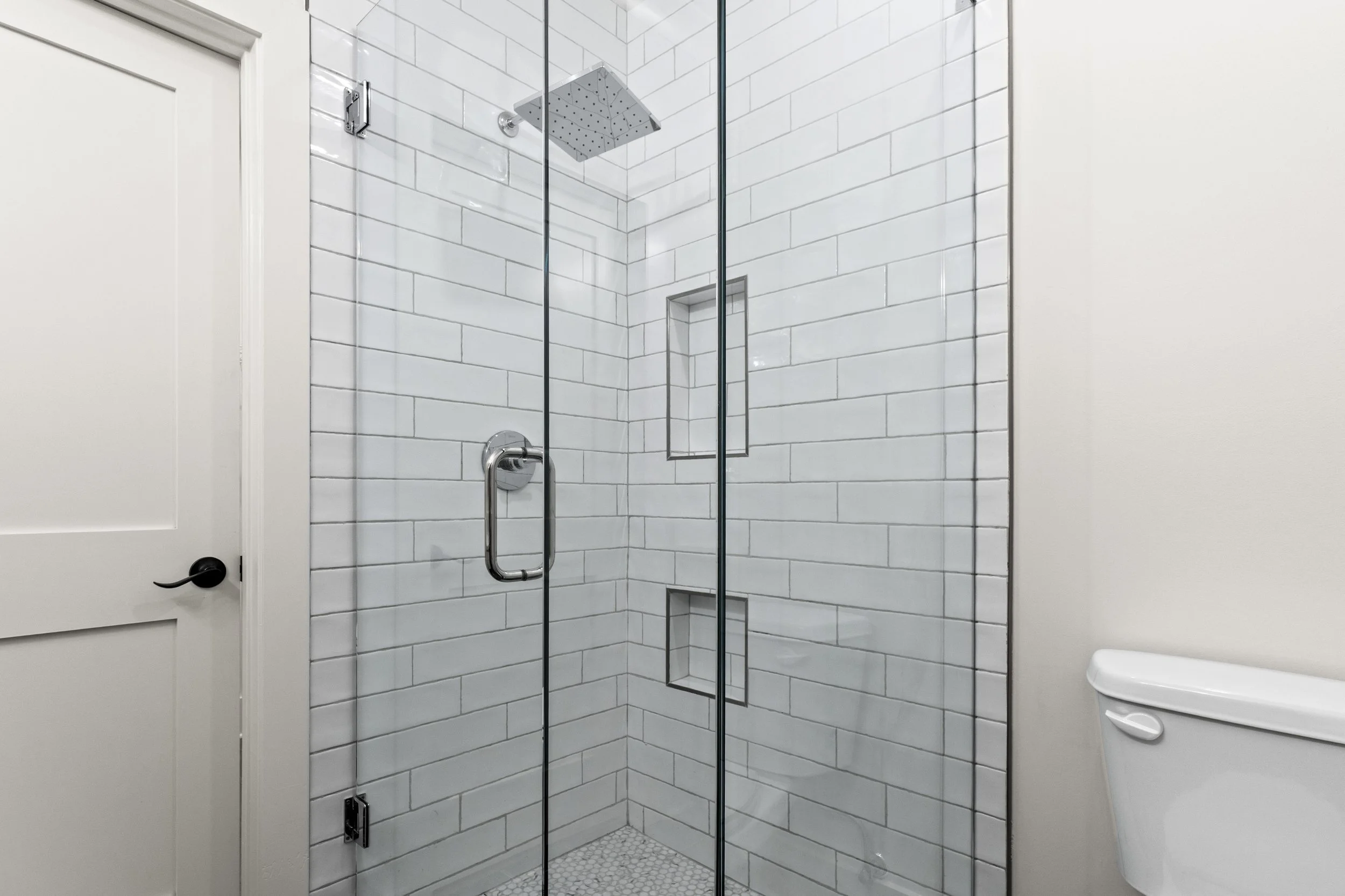 A modern glass-enclosed shower with white subway tile walls, a rain showerhead, and built-in shelving, next to a white door and partial view of a toilet.
