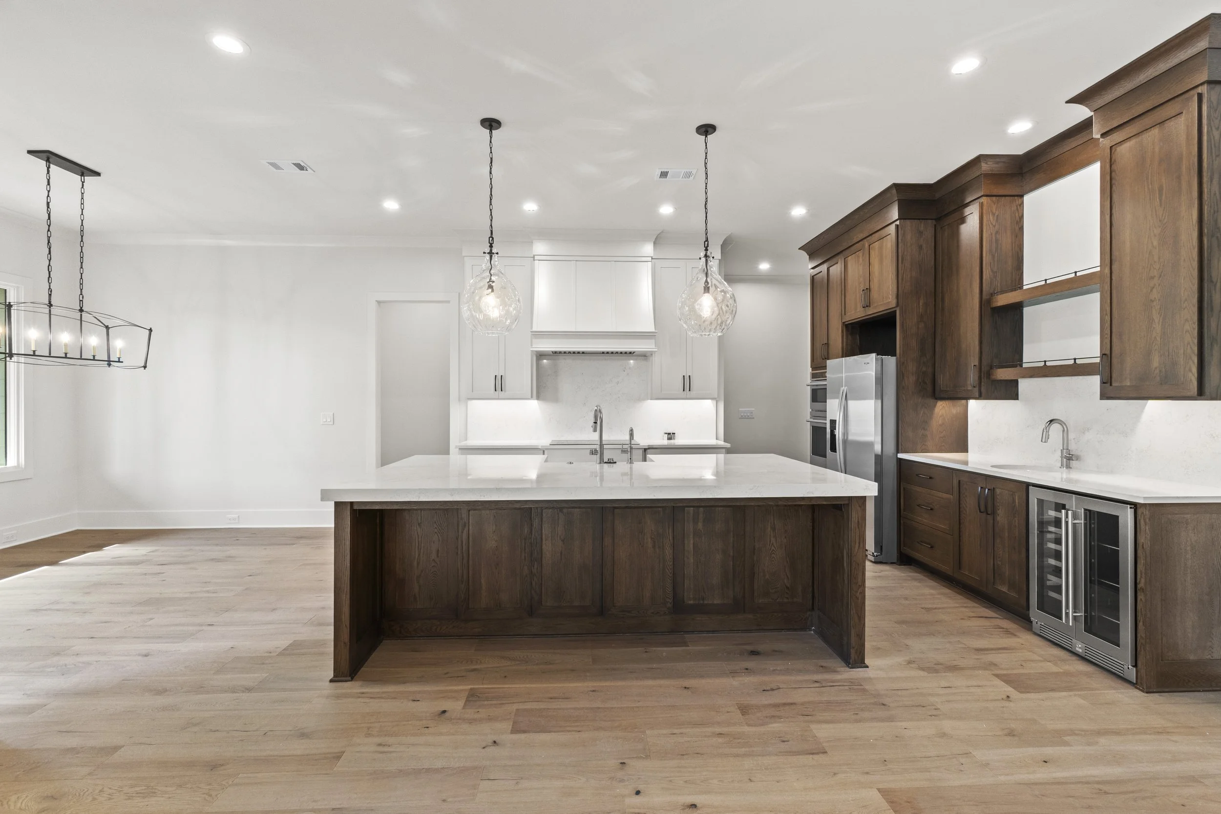 Modern kitchen with white walls, wooden flooring, and dark wood cabinetry. It features an island with a white countertop, stainless steel appliances, pendant lights, and recessed ceiling lights.