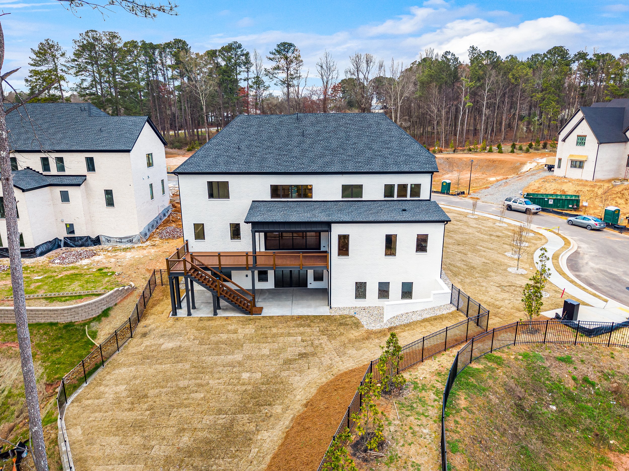 Aerial view of a residential area with multiple new white houses with dark roofs, a partly paved backyard with a wooden deck, young trees, and a surrounding wooded area.