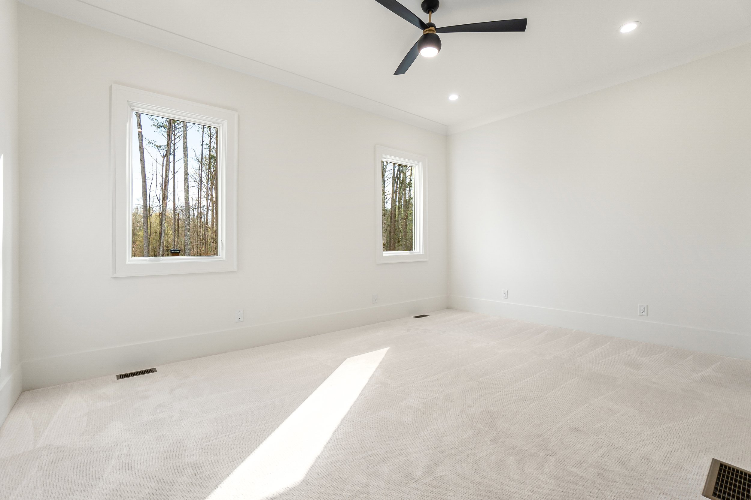 Empty room with white walls and ceiling, two windows showing trees outside, a ceiling fan with lights, and beige carpet flooring.