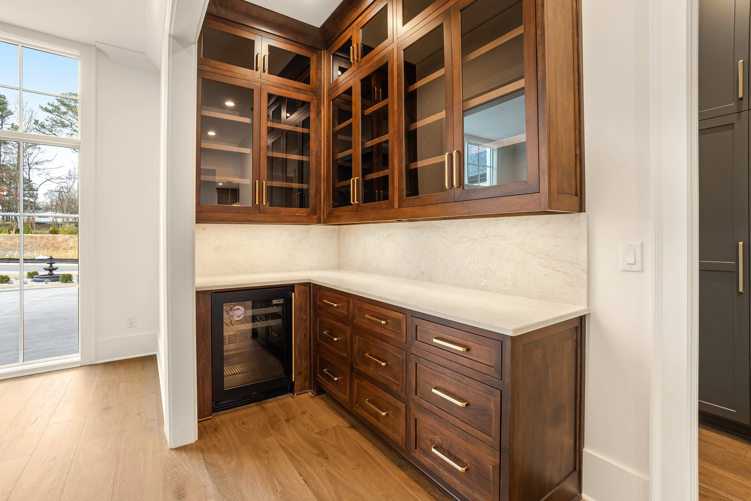 A corner of a kitchen with dark wooden cabinets, glass-paneled upper cabinets, a white countertop, and a wine fridge beneath the counter. There is a large window to the left showing an outdoor scene with trees and a parking lot.