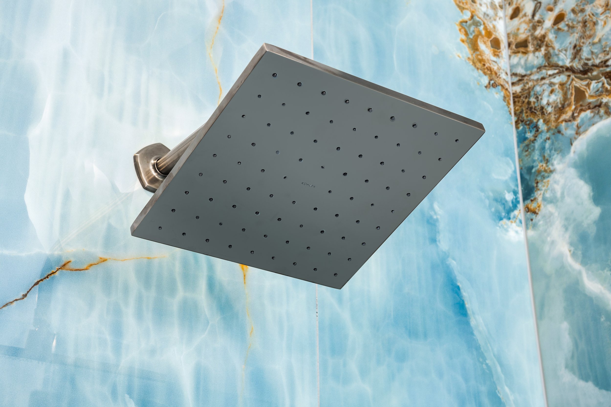 Close-up of a square ceiling showerhead with multiple small holes, mounted on a wall with blue and gold marble pattern.