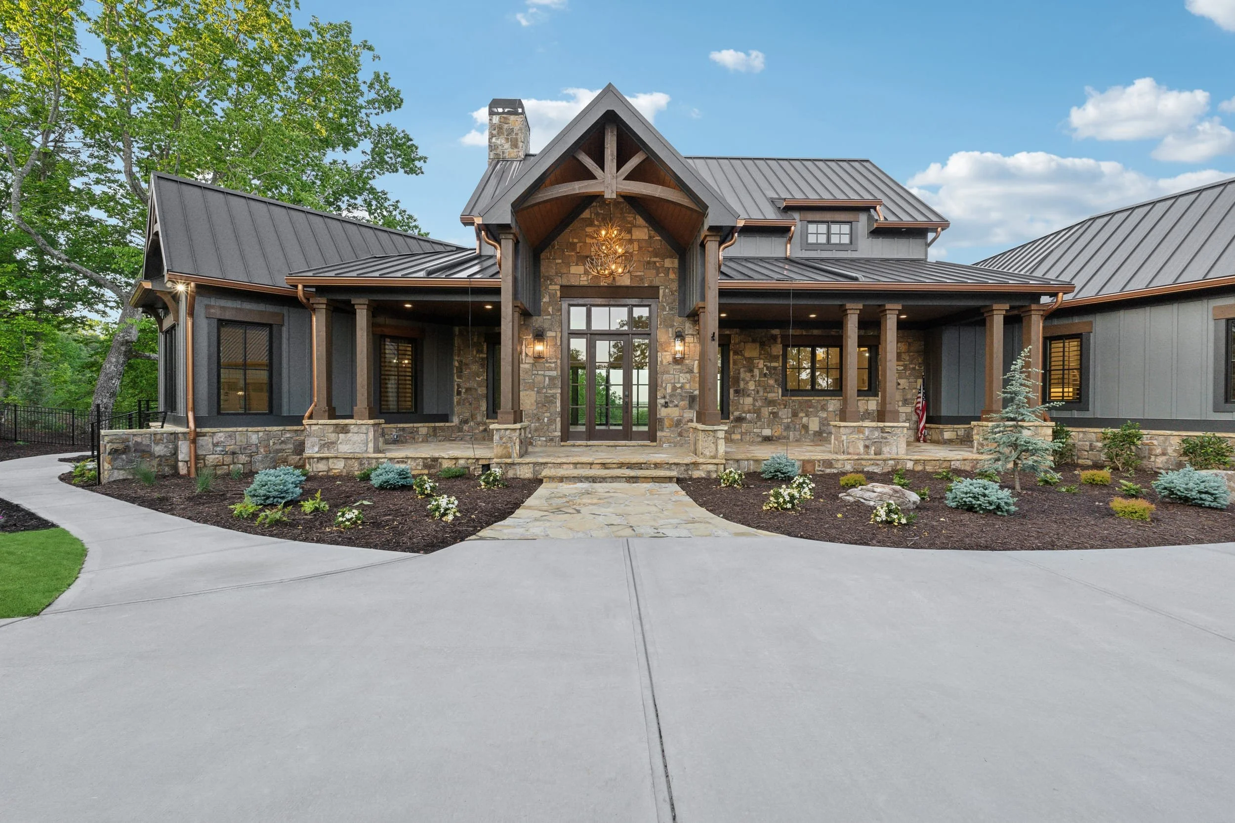 Front view of a large modern house with a stone and dark gray exterior, a metal roof, and a prominent entrance featuring a wooden porch with a chandelier, surrounded by a landscaped yard with small plants and a curved concrete driveway.