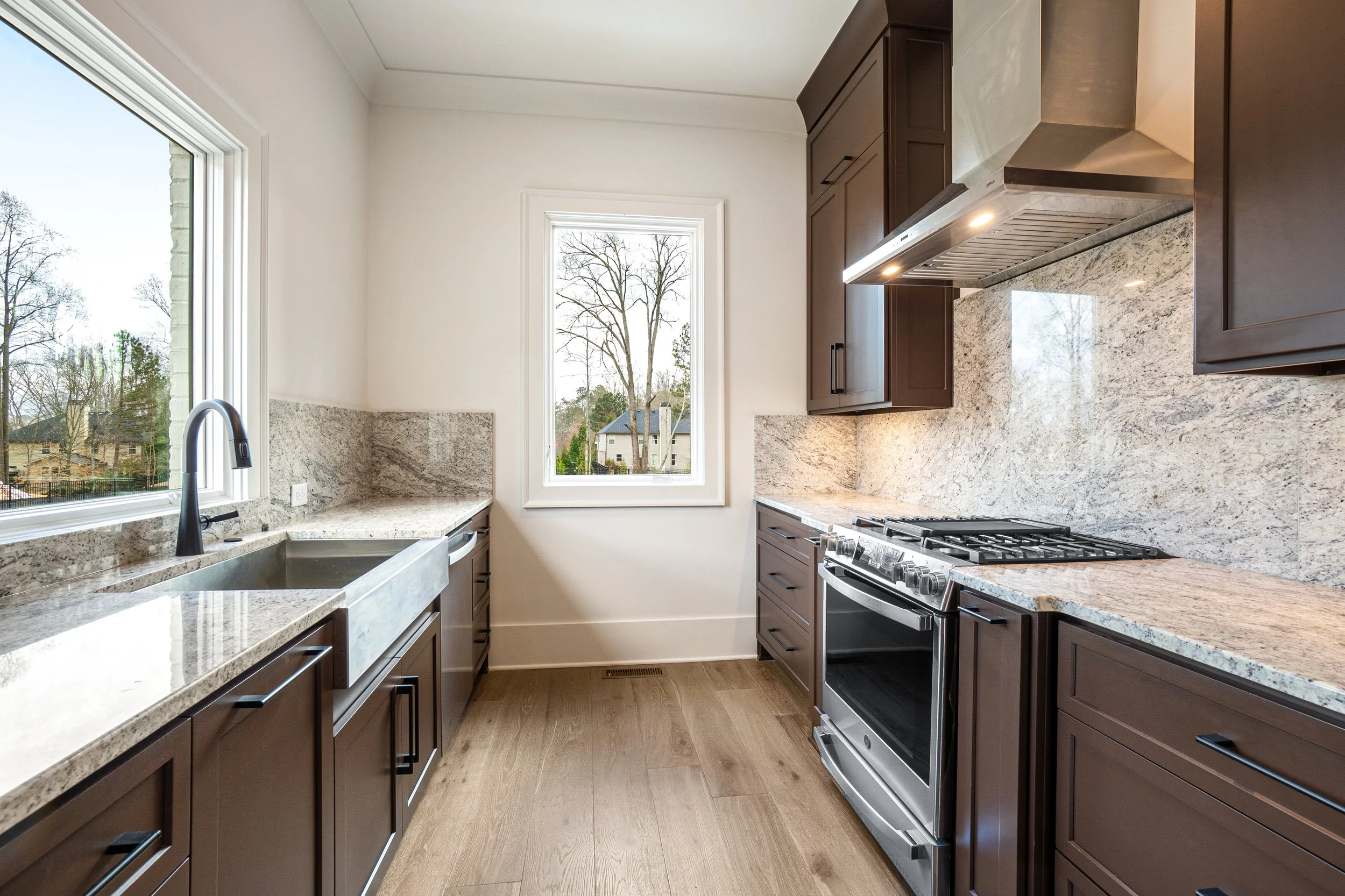 A modern kitchen with granite countertops and dark brown cabinets, a window showing trees outside, and stainless steel appliances including a stove and range hood.