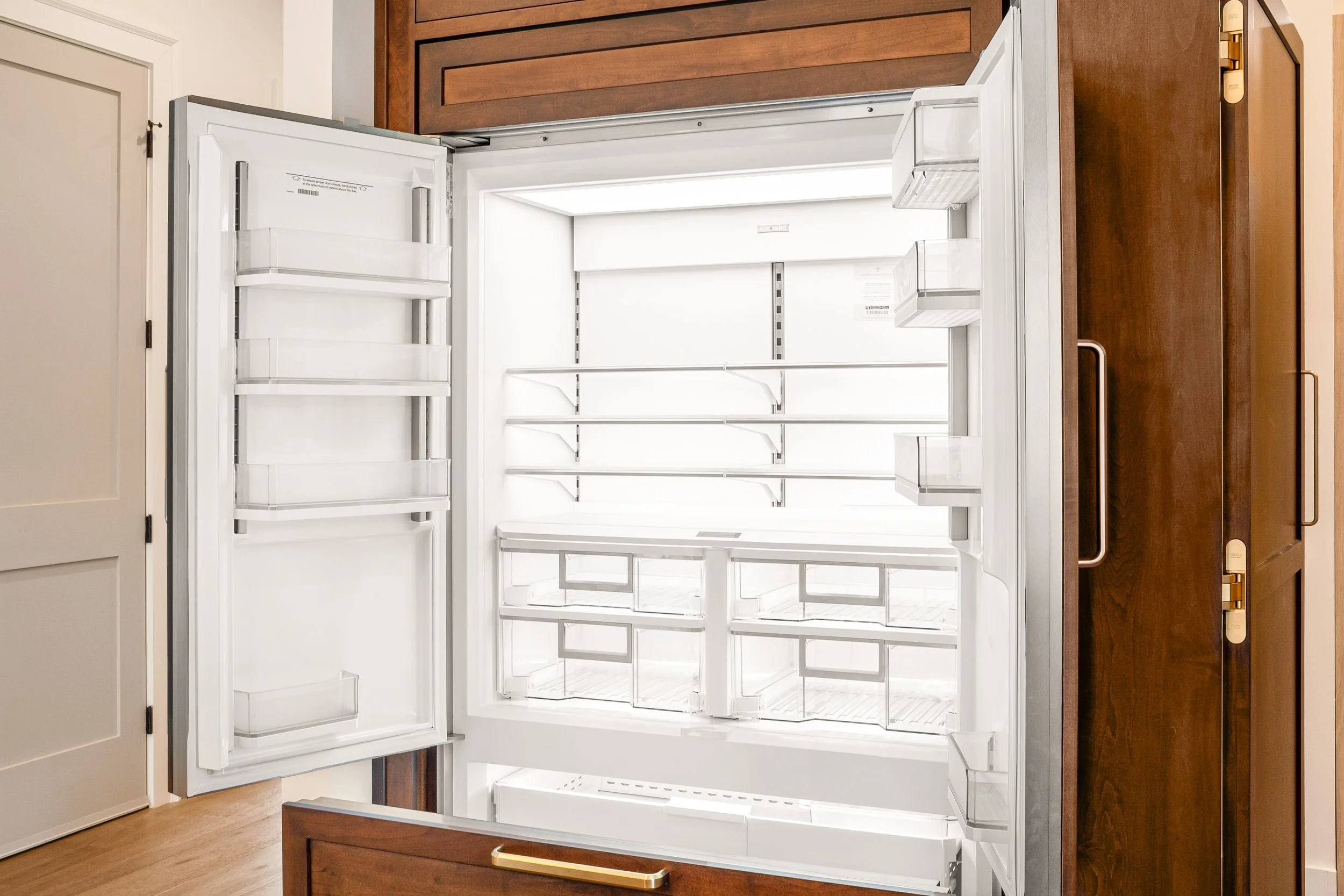 Empty white refrigerator with the door open in a kitchen, showing interior shelves and compartments.