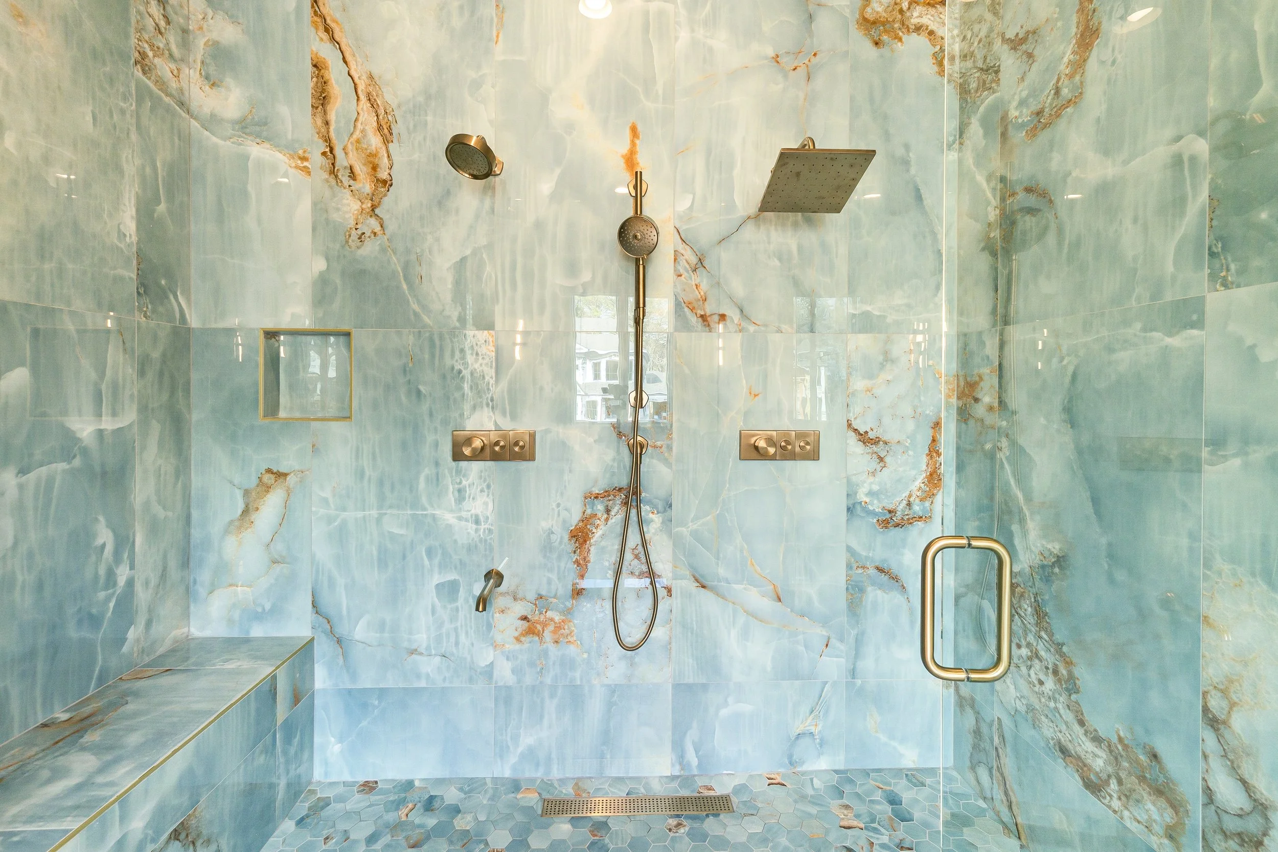 A modern shower with blue and brown marble tiles on the walls and floor, featuring a rain showerhead, a handheld showerhead, and a glass door with a metal handle.