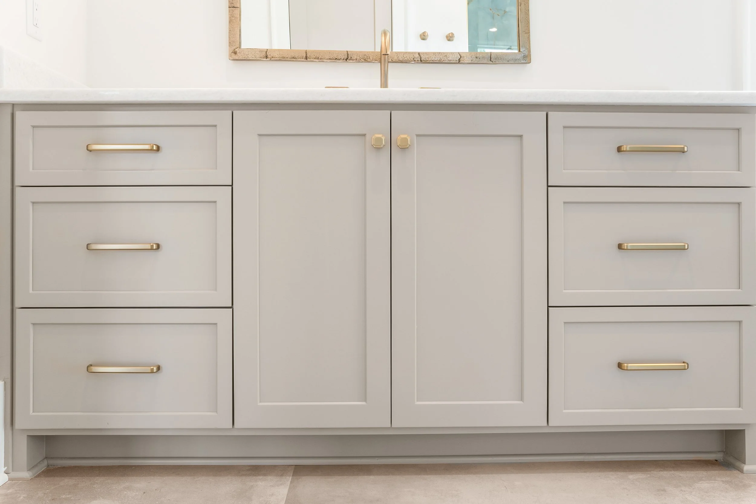 Light grey kitchen cabinet with gold handles below a mirror and white countertop with a sink faucet.