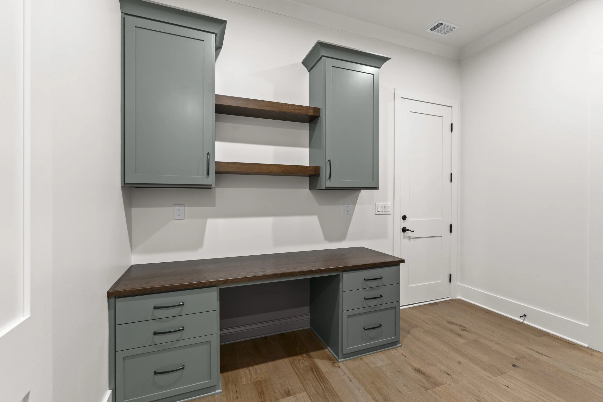 A corner of a room with built-in cabinets painted in light gray, with black handles, and open shelving with wood surfaces. There is a white door with black hardware on the right side, and the room has light wooden flooring and white walls.