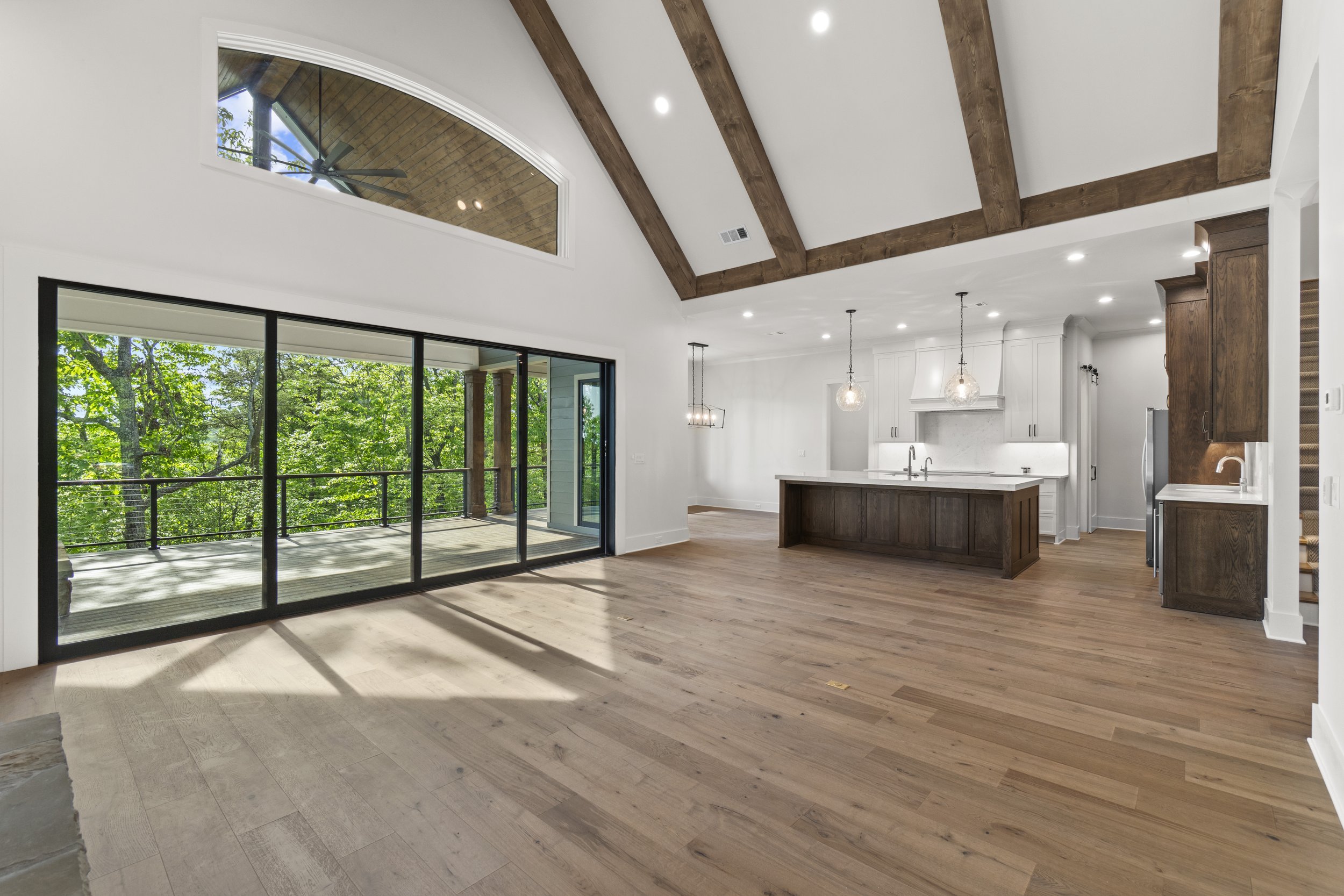 Interior of a bright modern living space with wooden vaulted ceiling, large sliding glass doors opening to a balcony with trees outside, and a kitchen with a white island and hanging pendant lights.