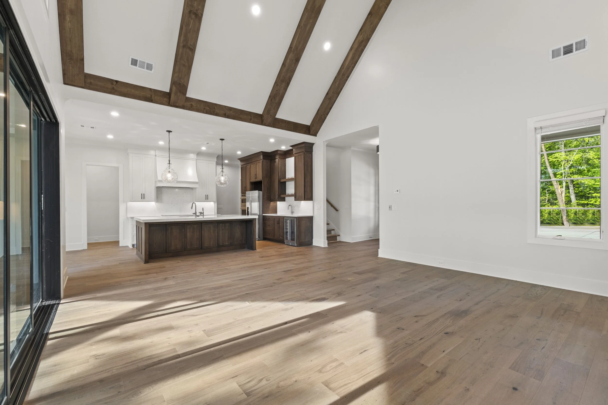 Open-concept living space with high vaulted ceiling supported by wooden beams, white walls, laminated wood flooring, large window with trees outside, kitchen with central island, cabinetry, and hanging pendant lights.