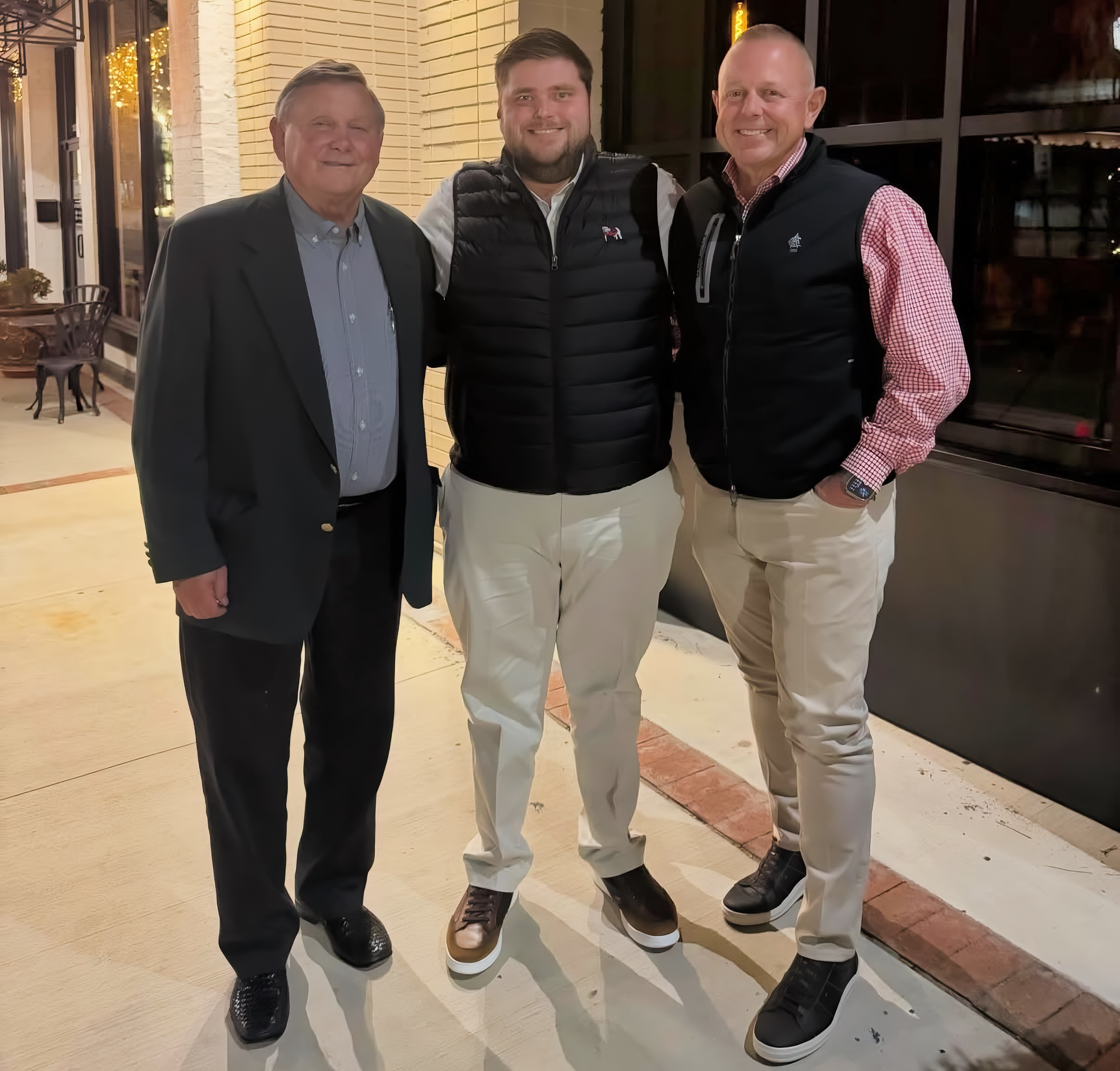 Three men standing side by side on a sidewalk outside a building at night, smiling, wearing casual and semi-formal attire, including vests and dress shirts.