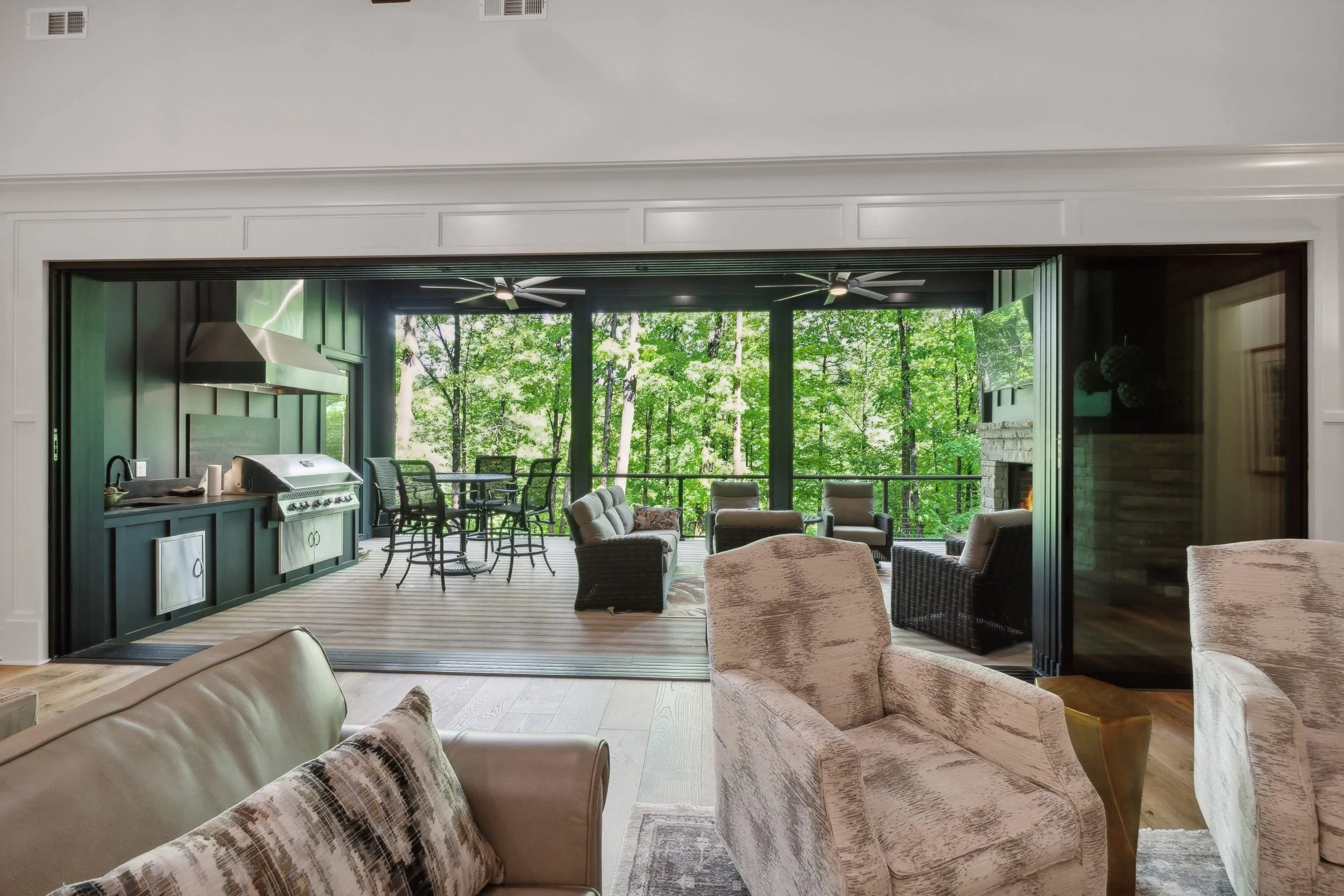 Indoor living space with a view of a wooded outdoor area, including ceiling fans, sofas, chairs, and a kitchen area.